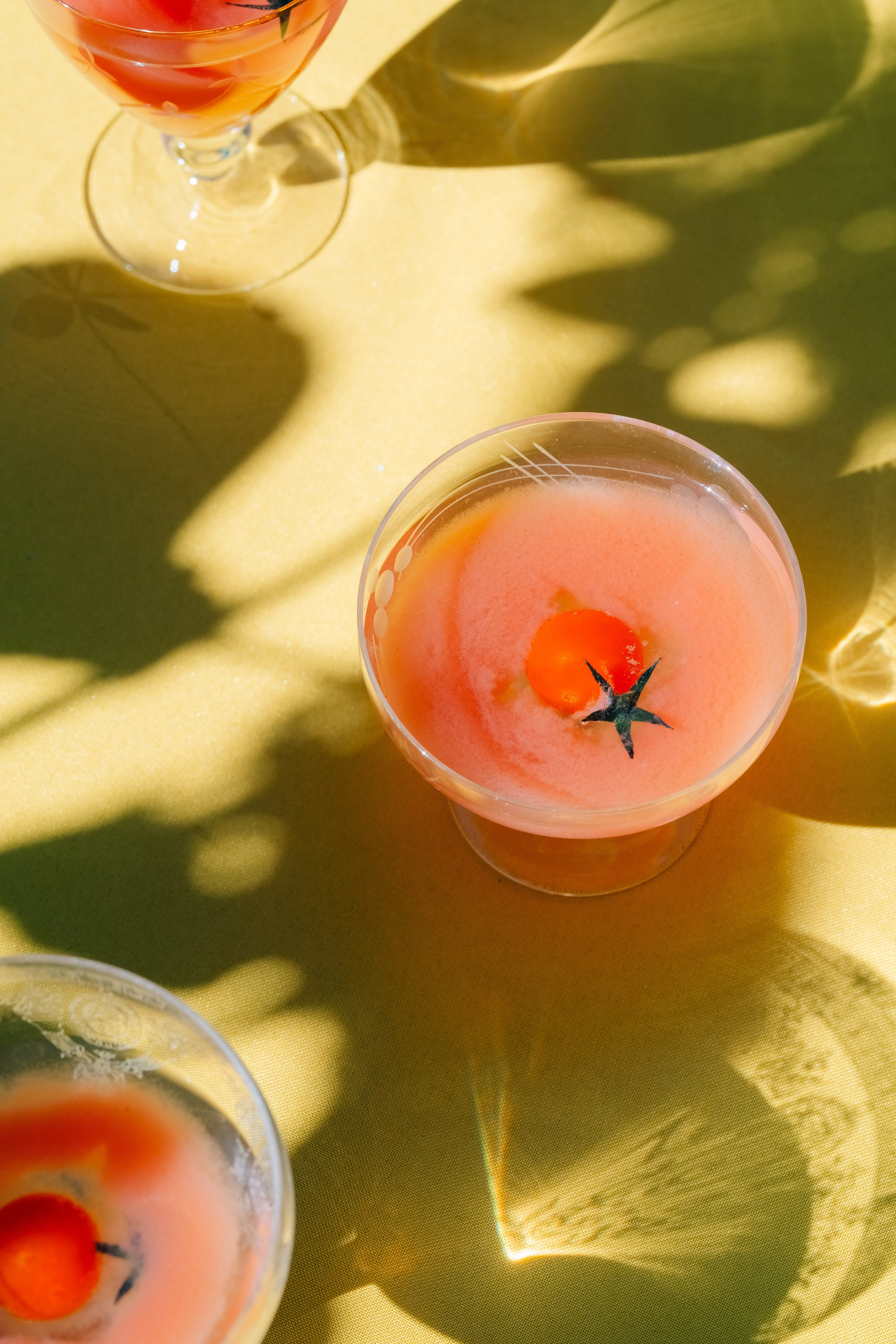 Cocktail de tomate servido em copos com luz e sombras naturais. Uma imagem vibrante de um cocktail de tomate servido em copos transparentes, com tomates cereja a flutuar no topo, sobre um fundo amarelo com sombras divertidas e reflexos de luz.