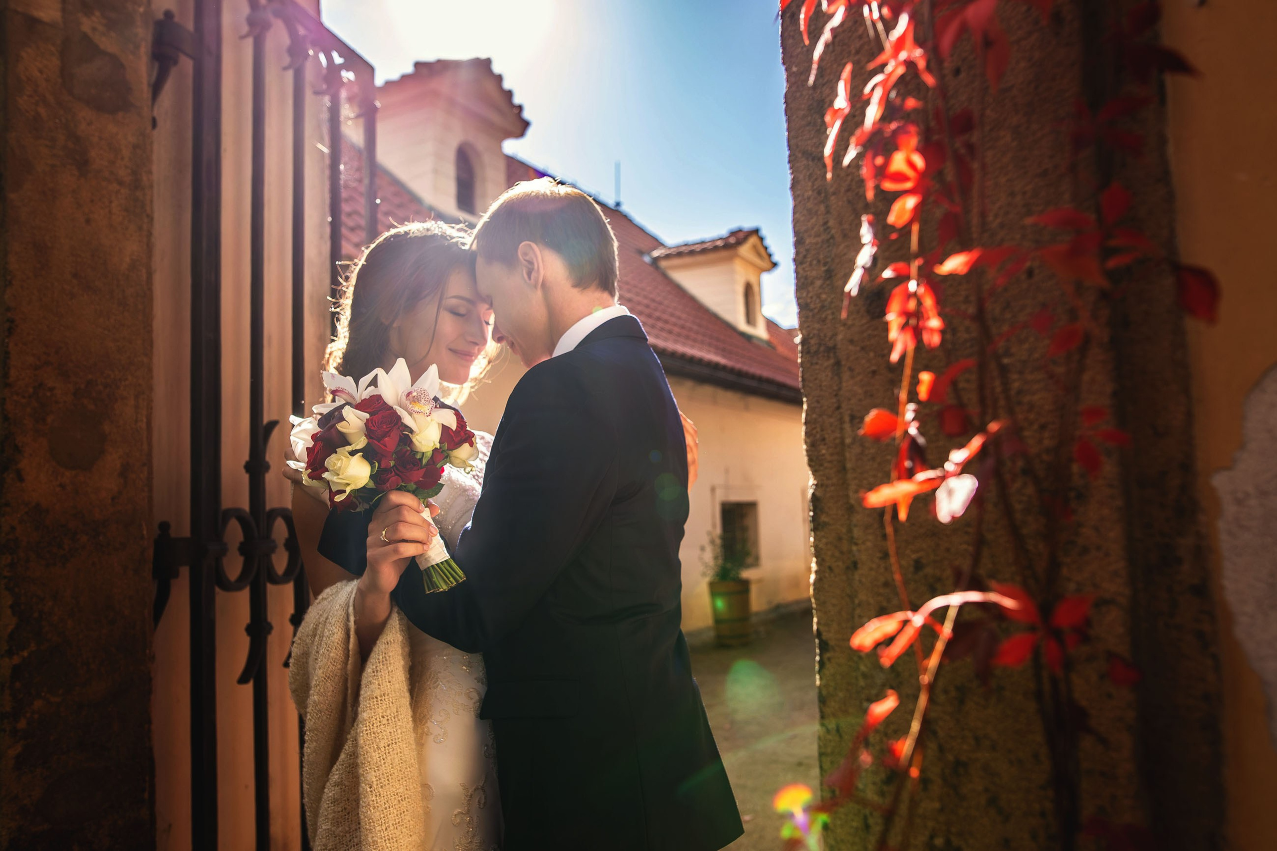Brunette bride embraced by groom in sun-flared doorway to historic garden, bouquet near face.