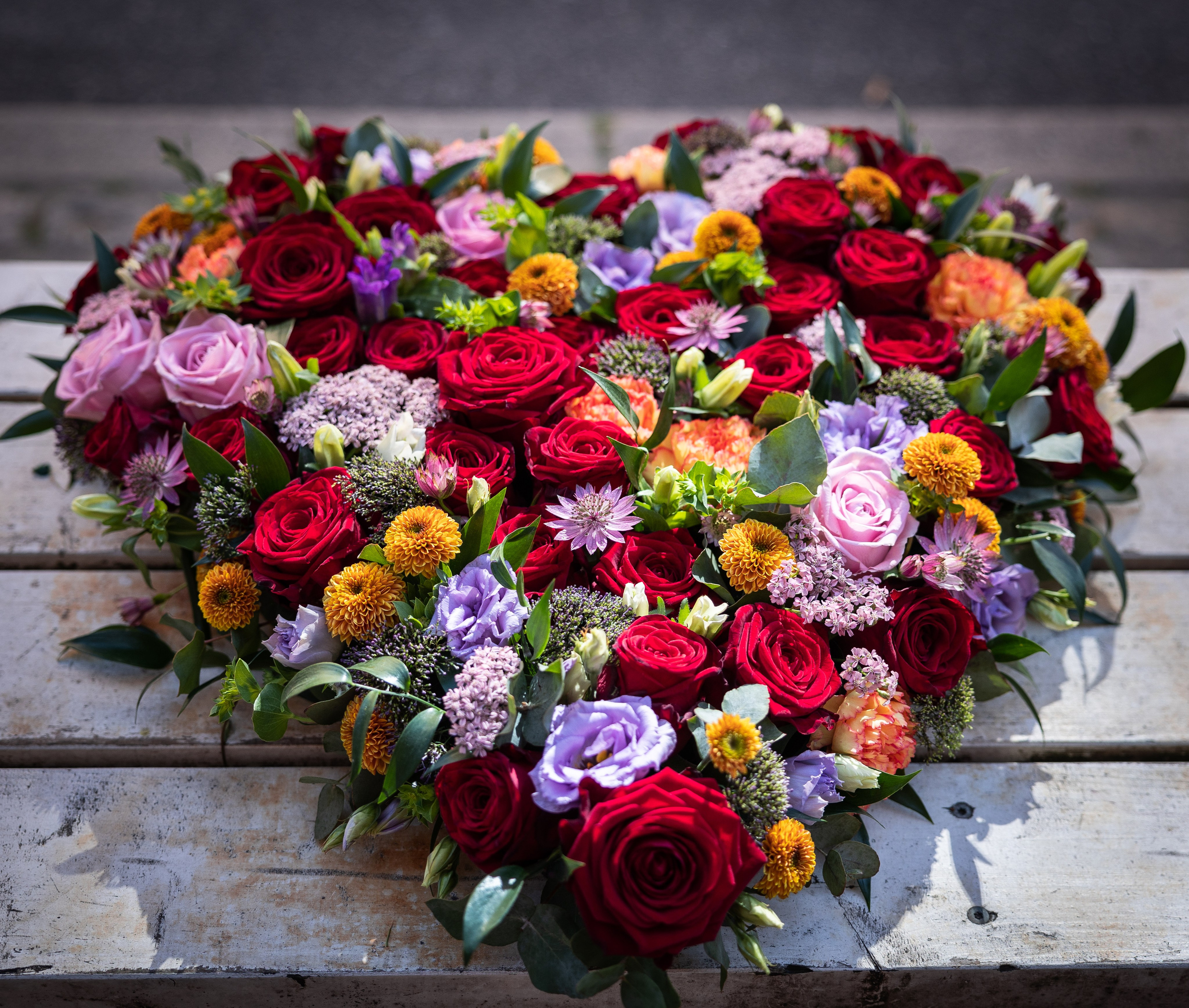 Hjerte, hvor blomsterne sidder tæt sammen i blå, orange og rødlige toner. Små og store blomster er kombineret i et flot sammenspil, som giver et levende udtryk. En sidste kærlig hilsen, som tager sig smukt ud på kirkegulvet.