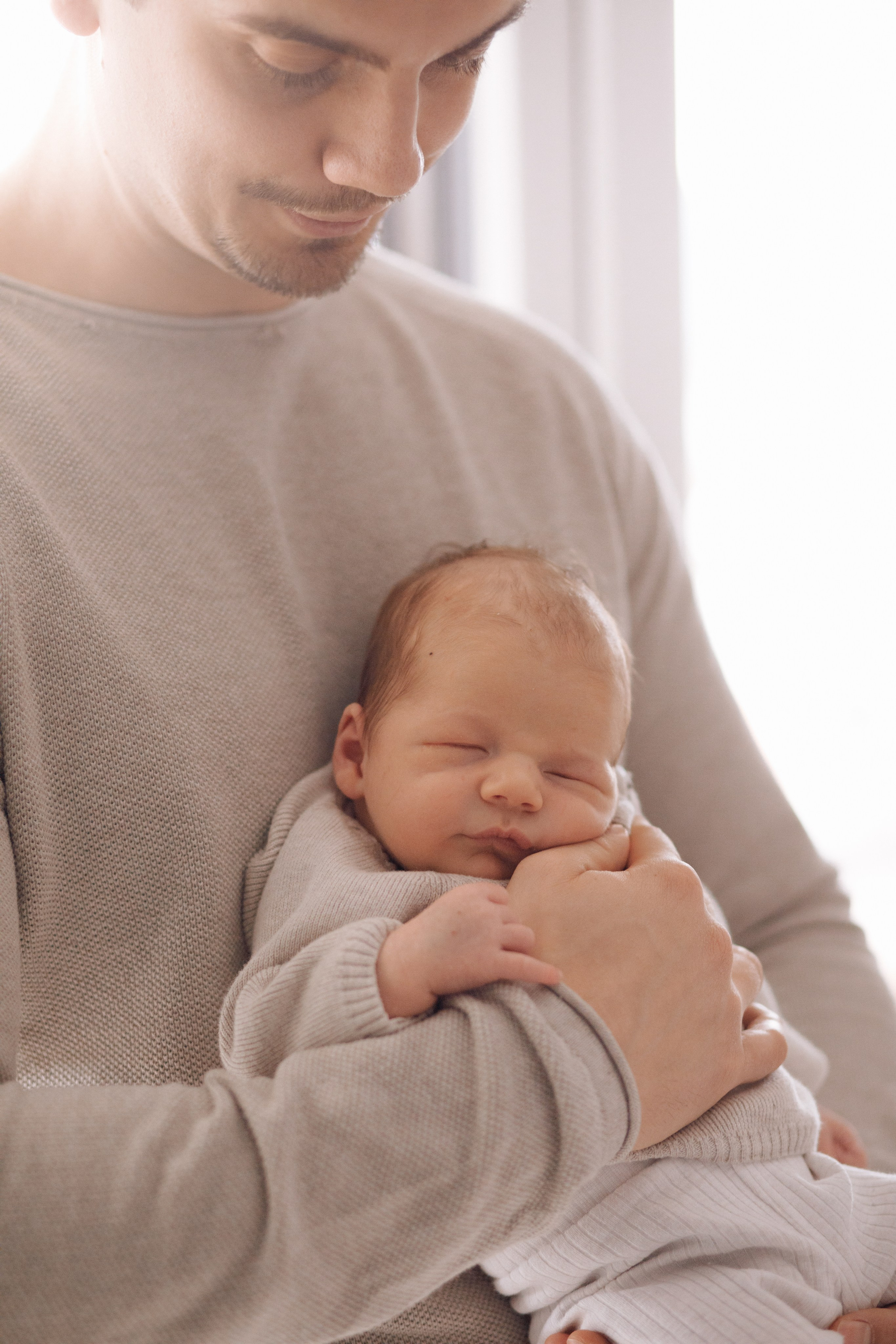 Papa umarmt sein Neugeborenes in einem lichtdurchfluteten Raum während eines emotionalen Fotoshootings.