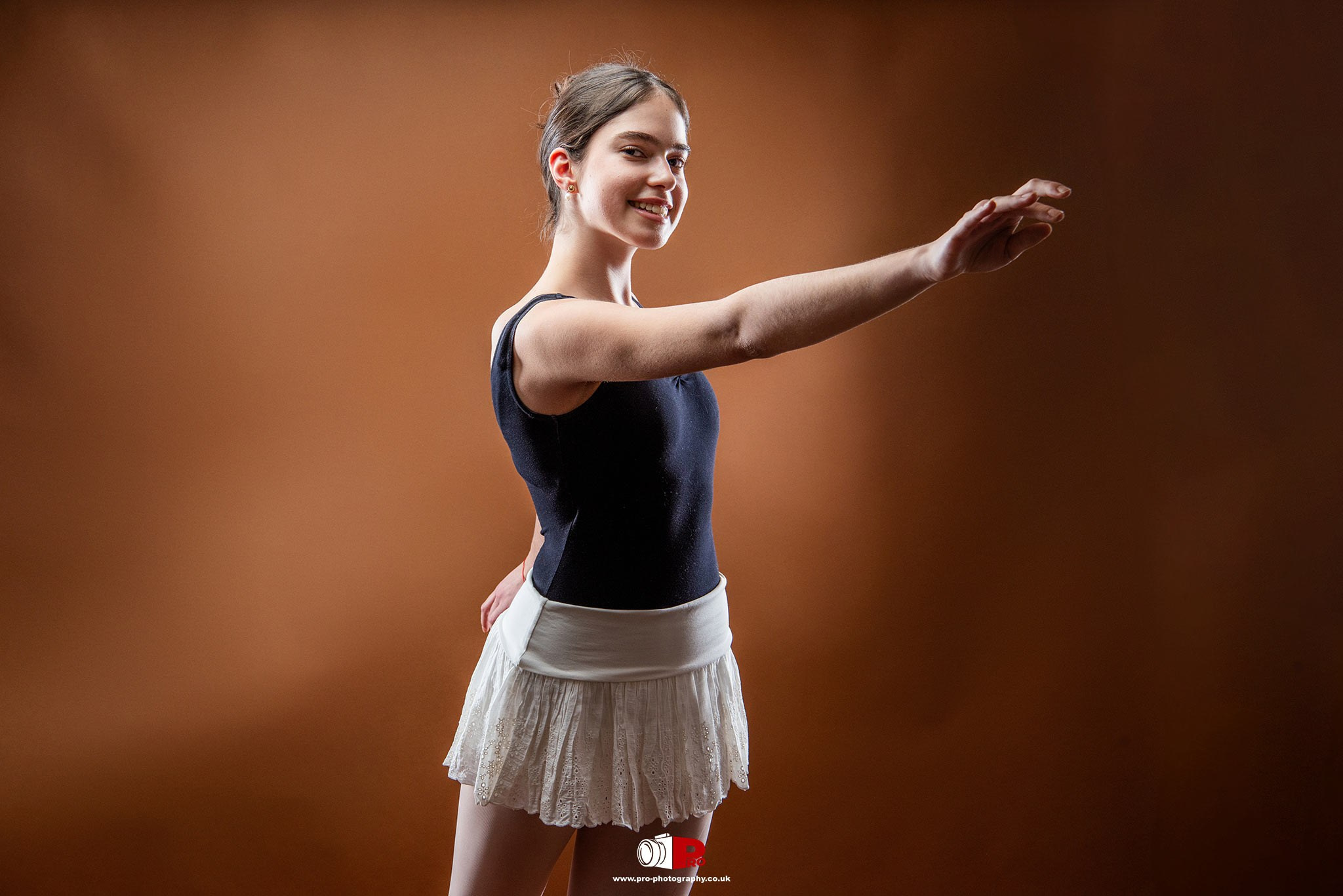 Young ballet dancer posing gracefully in a black leotard and white skirt against a warm brown backdrop.