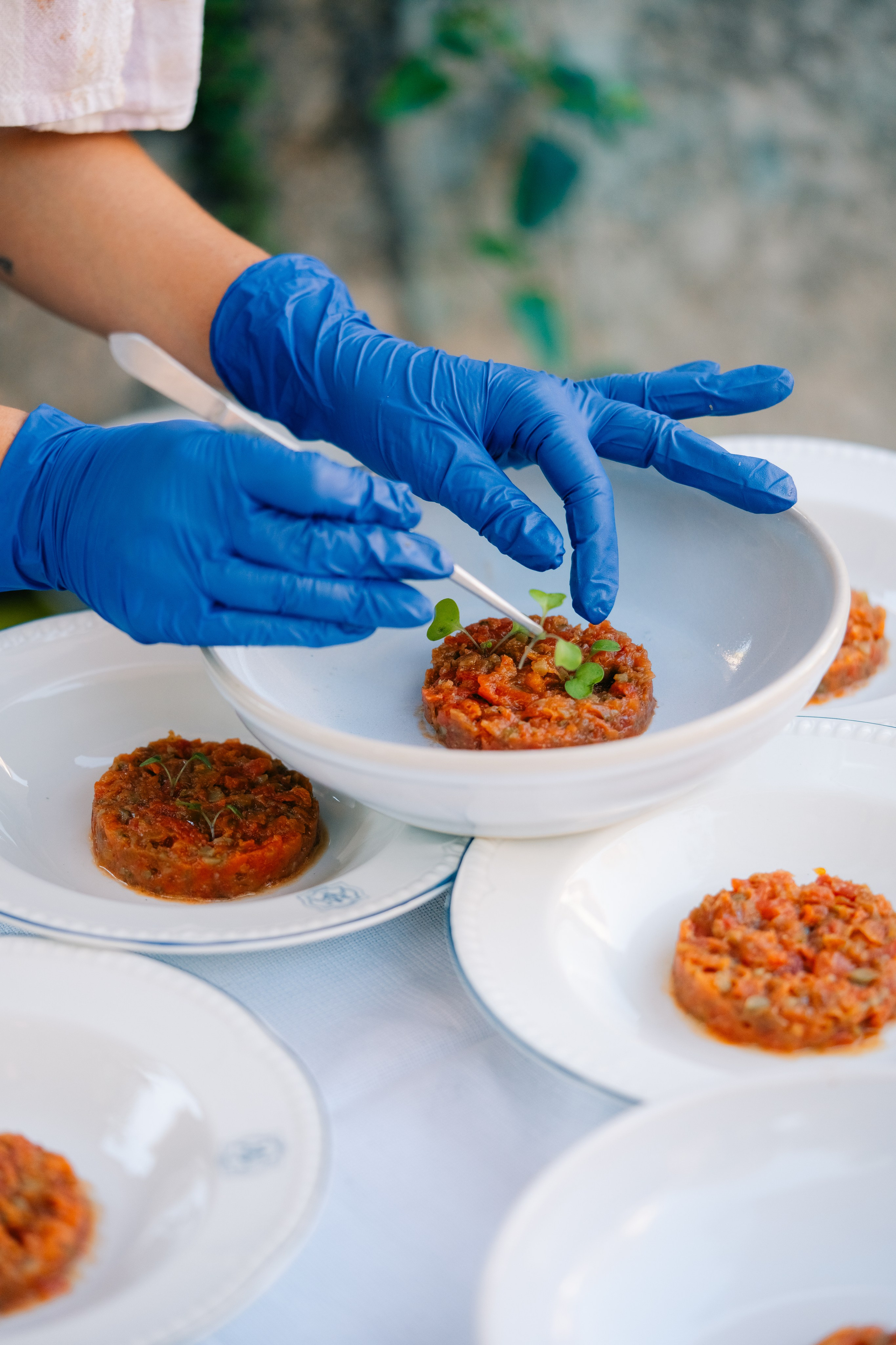 Chef a chapear tártaro vegetal com luvas azuis. Um chef que usa luvas azuis prepara cuidadosamente tártaro de vegetais em pratos brancos, adornando com microgreens para uma apresentação gourmet num ambiente ao ar livre.