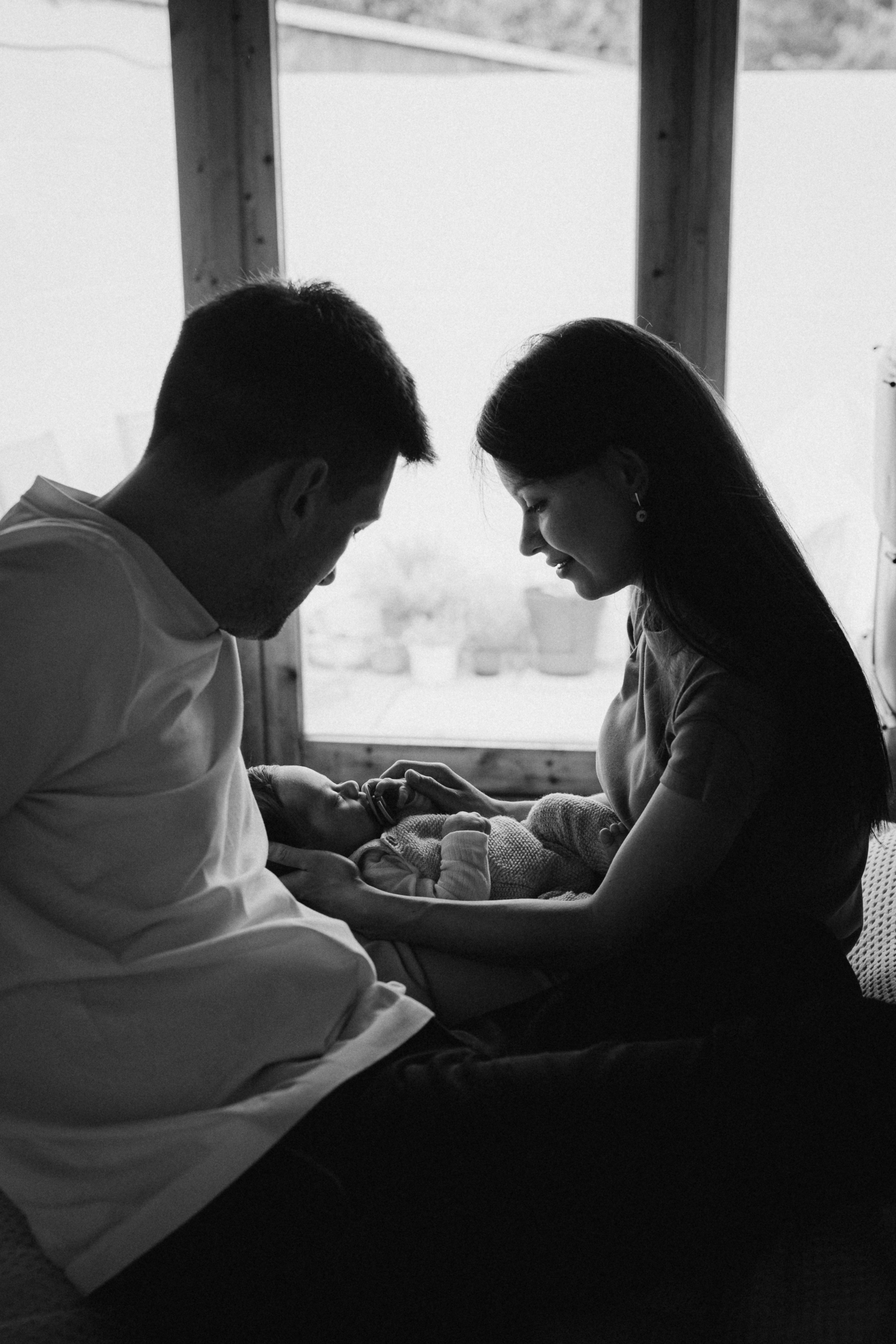 black and white photo of the parents hold their baby opposite the window