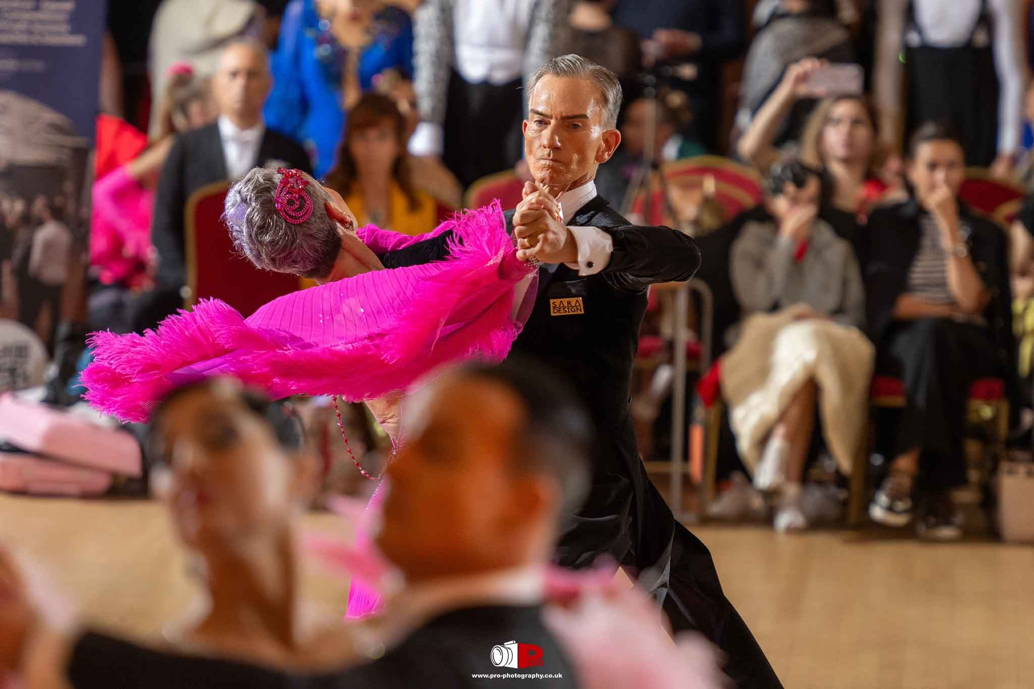Senior male and female ballroom dancers gracefully perform with vibrant pink feather accents.