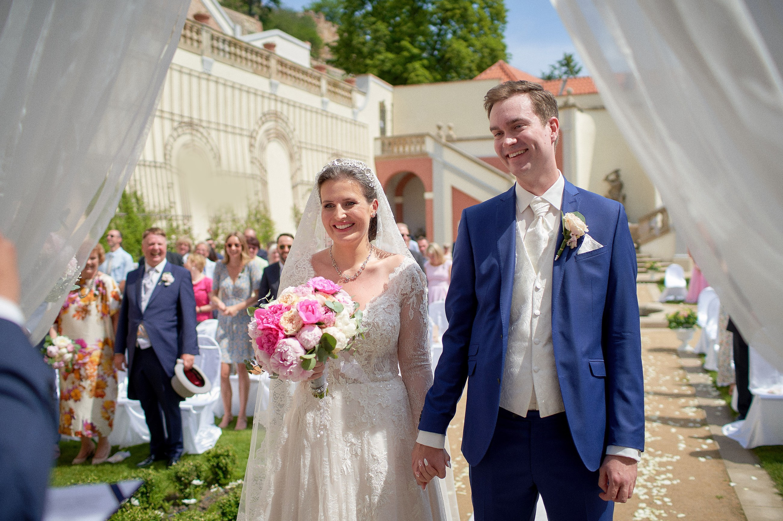 The smiling bride & groom are seen under the floral arch as their wedding guests look on from behind during their Ledebour Garden wedding.