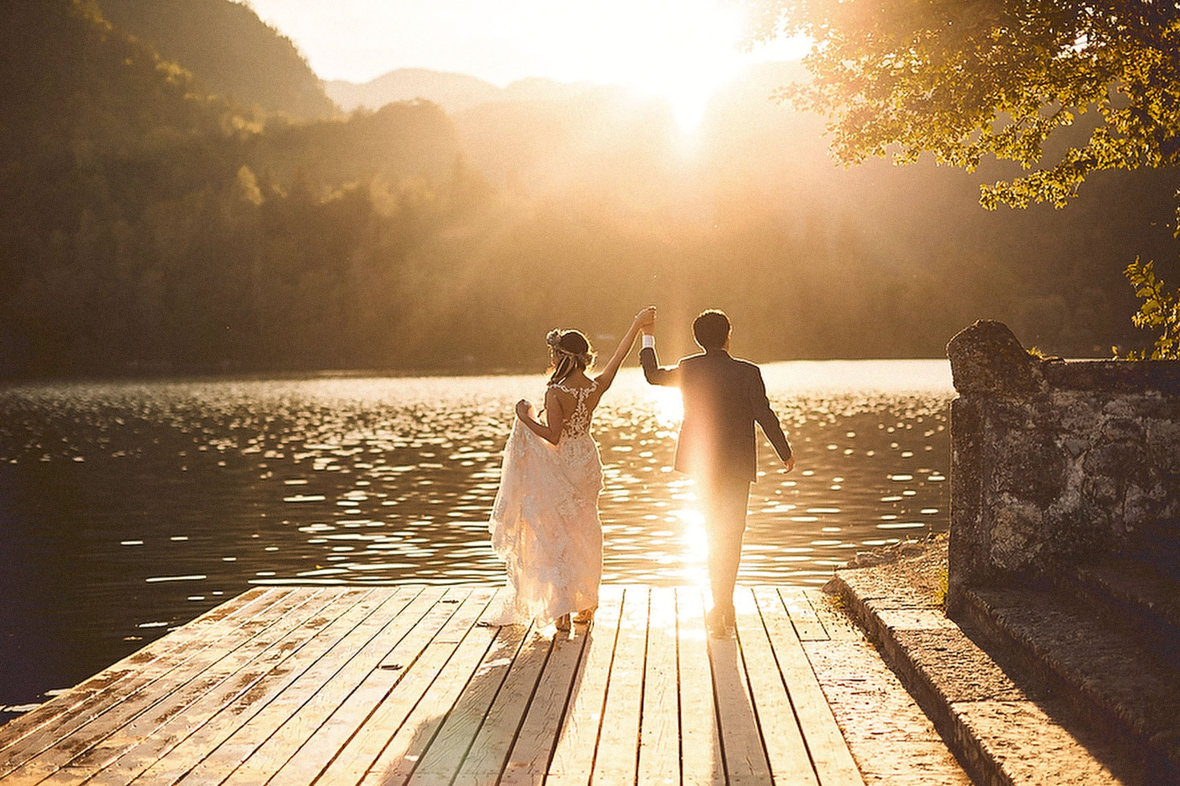 Japanese bride and groom celebrating with hands raised at Lake Bled, Slovenia, under spectacular sunlight."
