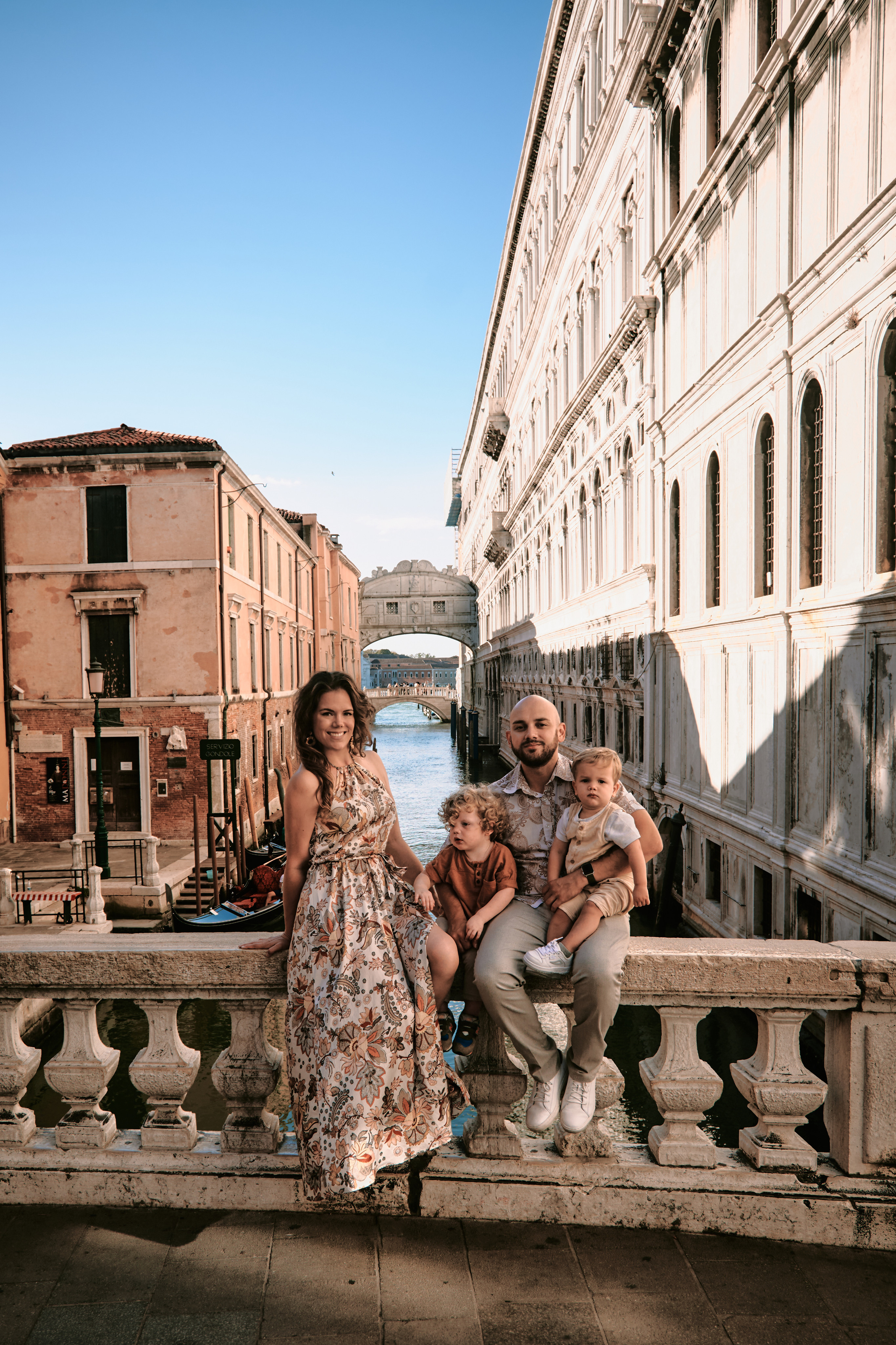 A happy family of four stands in front of the iconic St. Mark's Square, with the beautiful Venetian architecture and gondolas in the background.