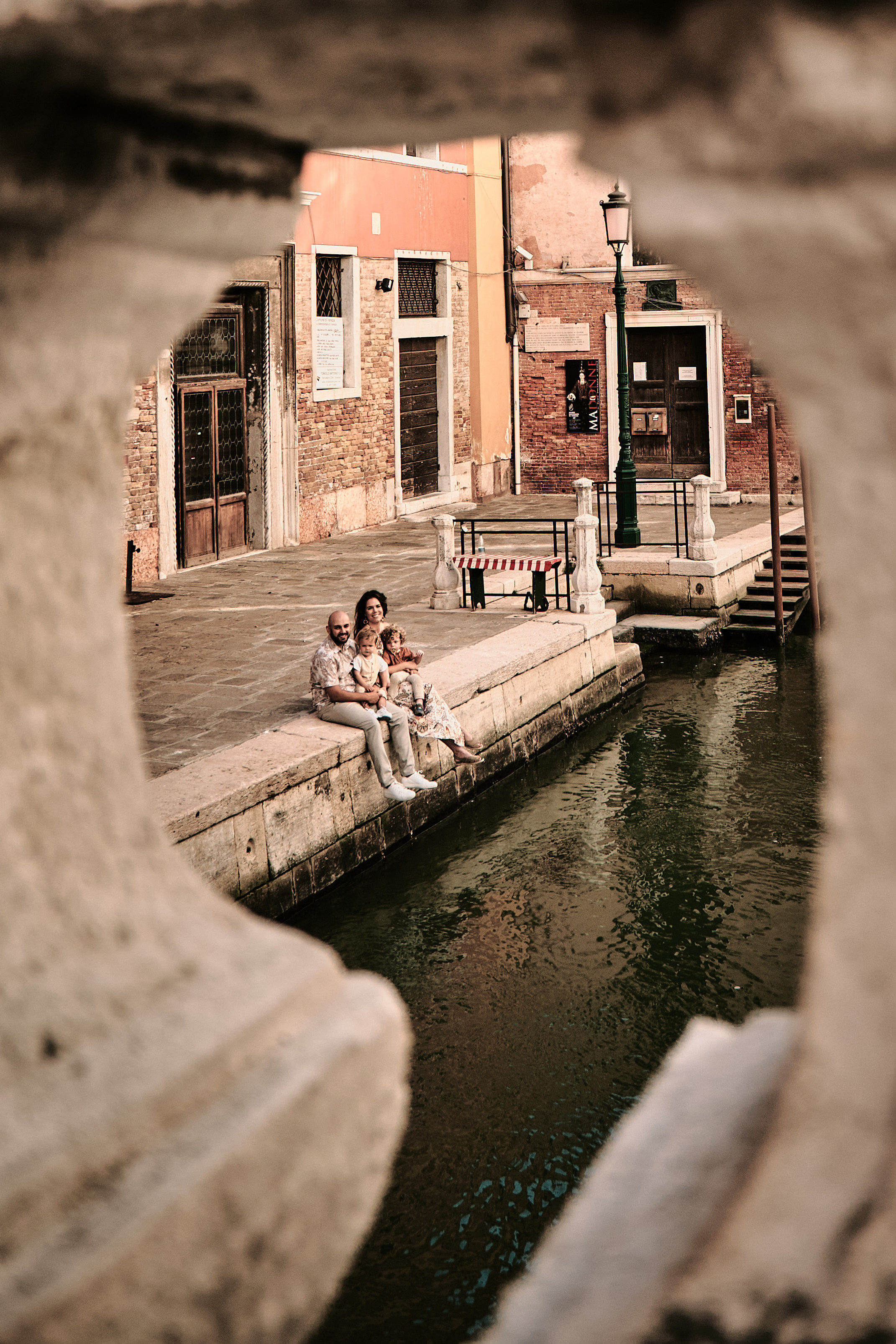 Two adorable children smile as they pose with their parents in a charming Venetian alleyway, surrounded by colorful buildings and flower pots.