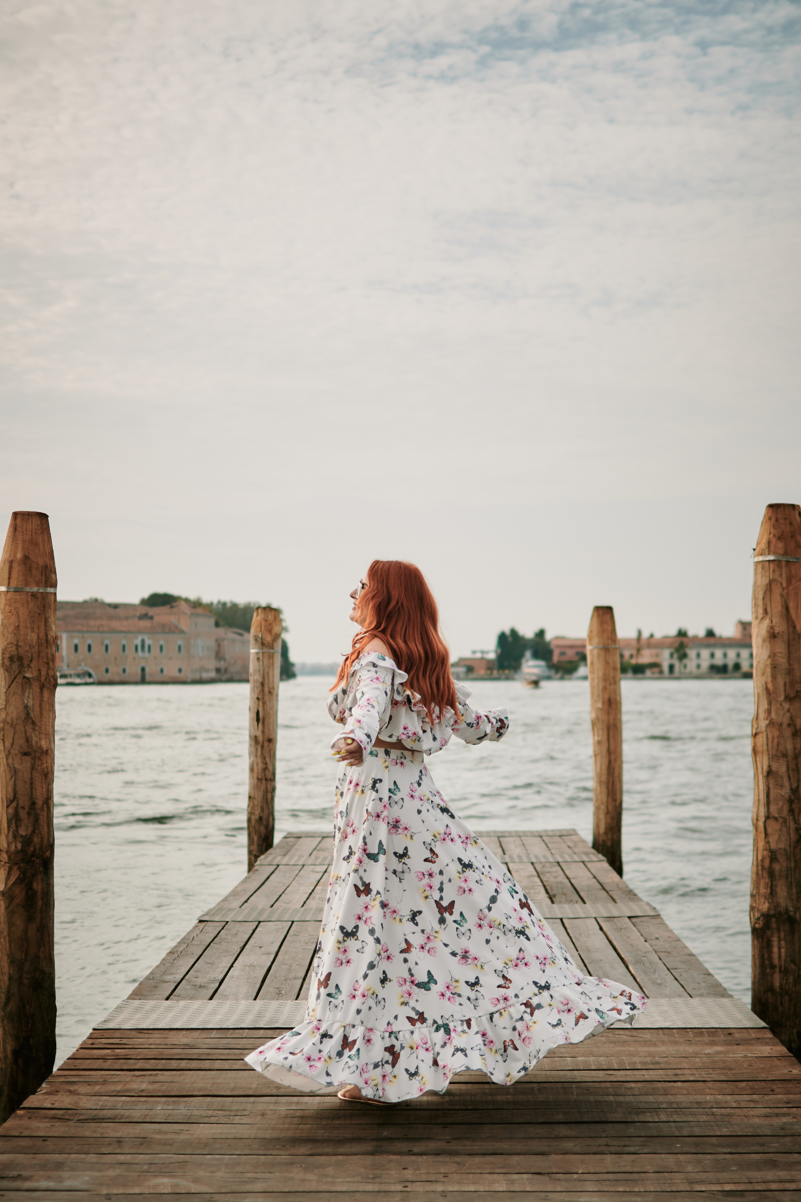 A young woman stands on laguna of Venice. T She wears a flowing dress that seems to dance in the breeze, accentuating her graceful movements. The intricate details of the nearby buildings and the vibrant colors of the street performers create a romantic and captivating atmosphere around her, making for a beautiful and memorable portrait.
