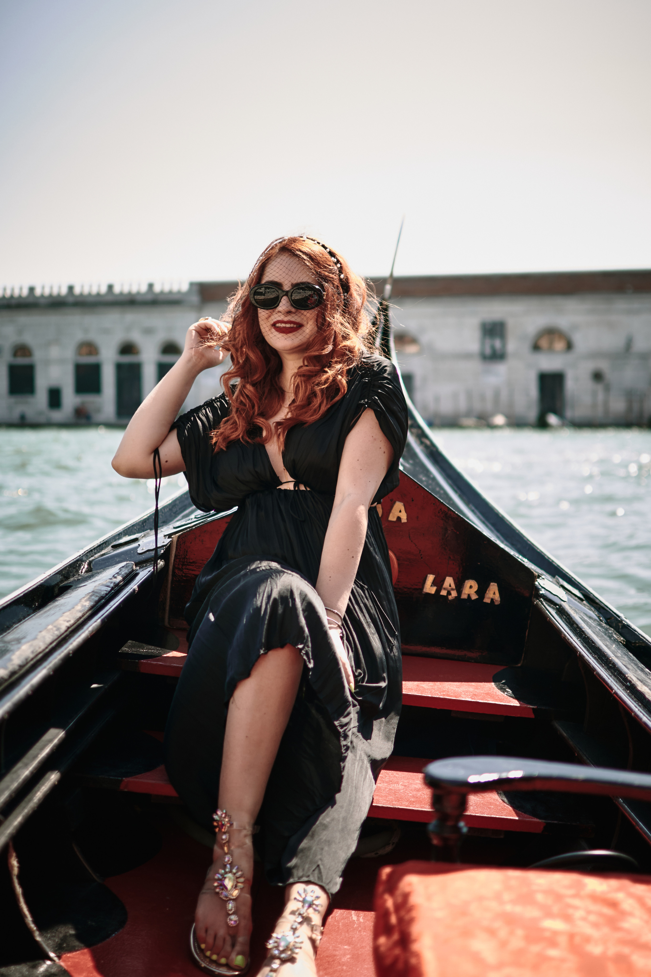 A young woman in a black dress sits on a gondola as it glides through the narrow streets of Venice. She gazes off into the distance, lost in thought, as the sunlight dances on the water around her. The picturesque buildings of the city rise up on either side, their pastel colors and ornate details adding to the romantic atmosphere. The woman's elegant dress and captivating presence seem perfectly at home in this enchanting setting, creating a sense of mystery and allure that draws the viewer in.