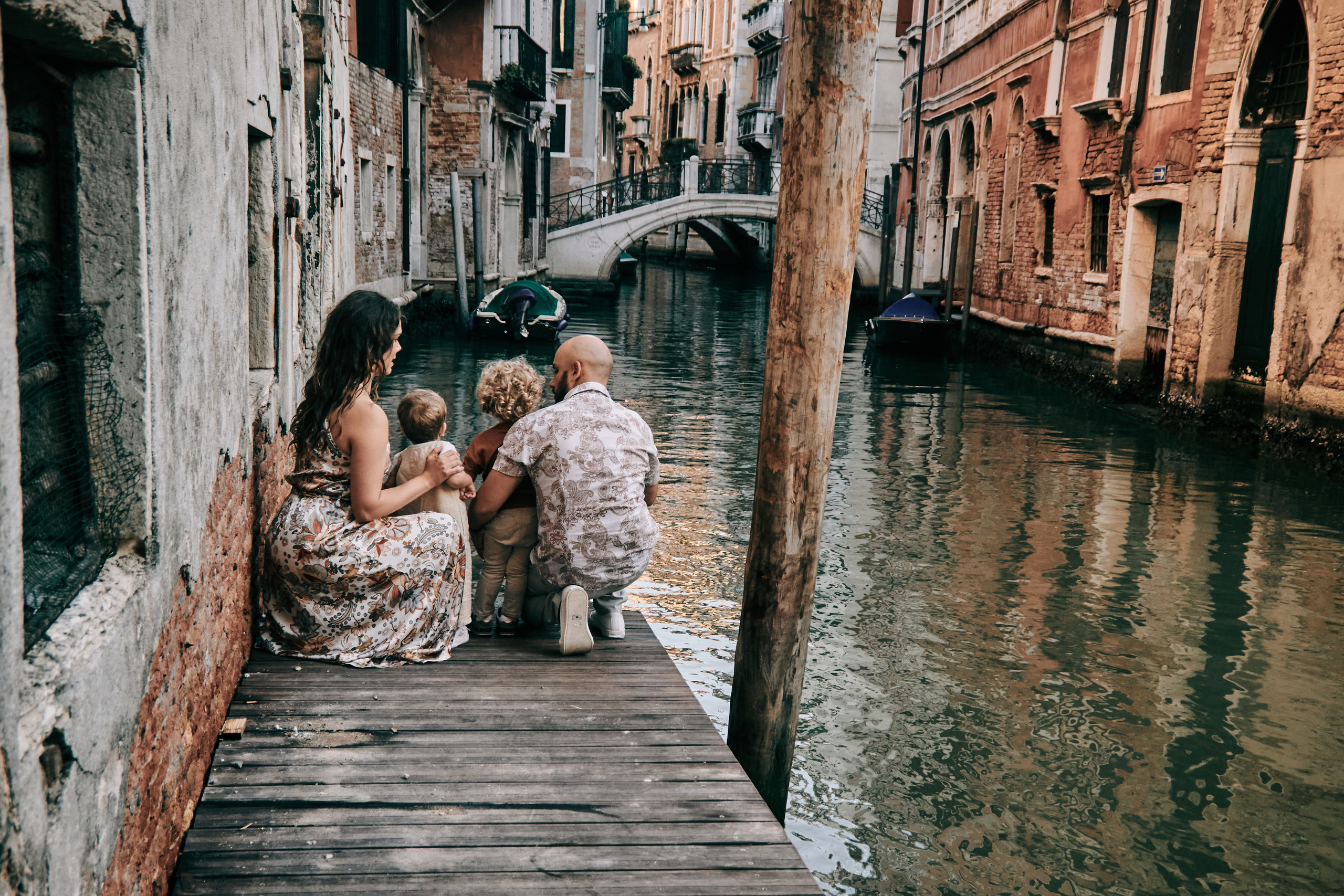 A happy family of four stands in front of the iconic St. Mark's Square, with the beautiful Venetian architecture and gondolas in the background.