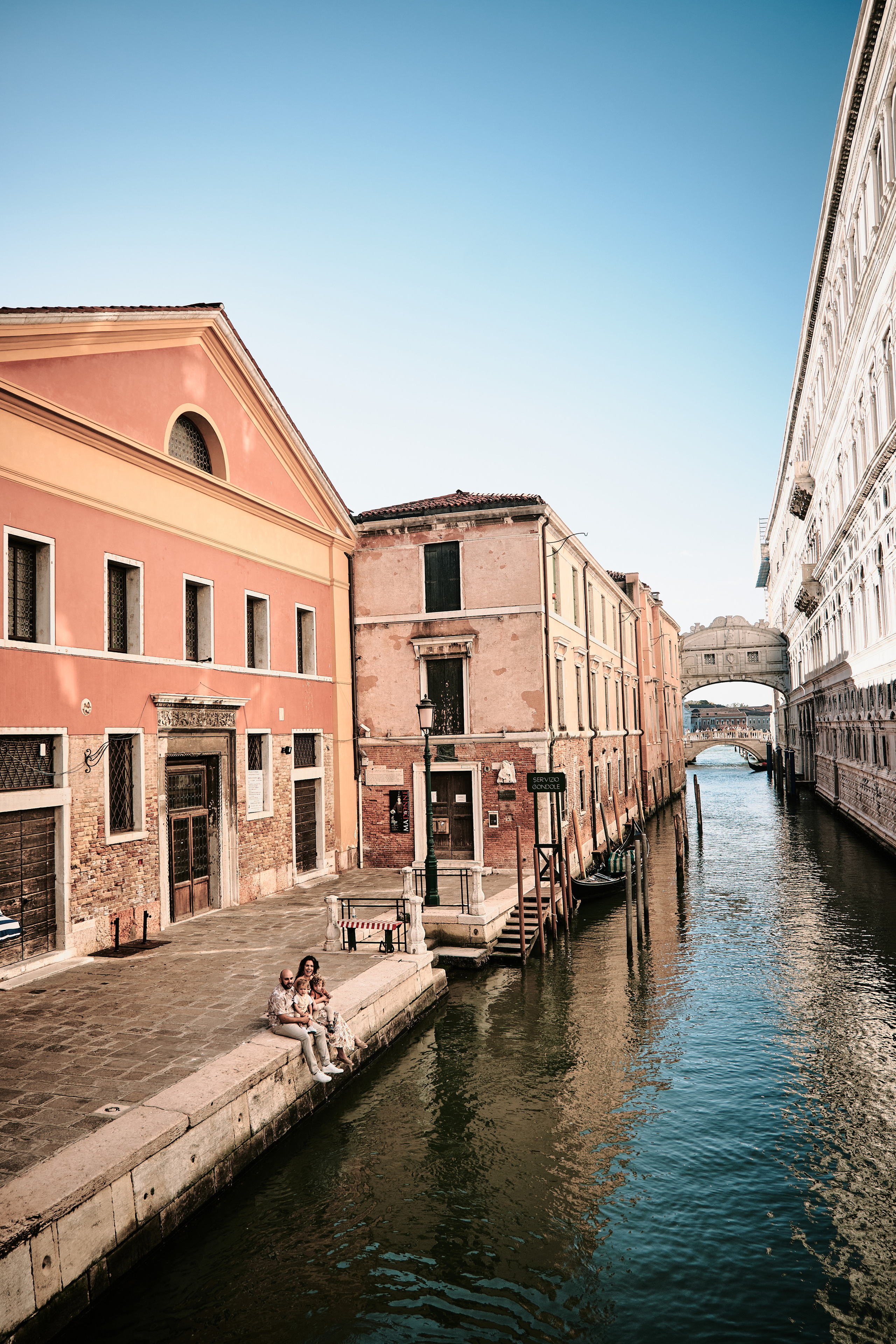 A loving family takes a stroll by the  Canal, with the stunning Bridge of Sighs and gondolas passing by in the background