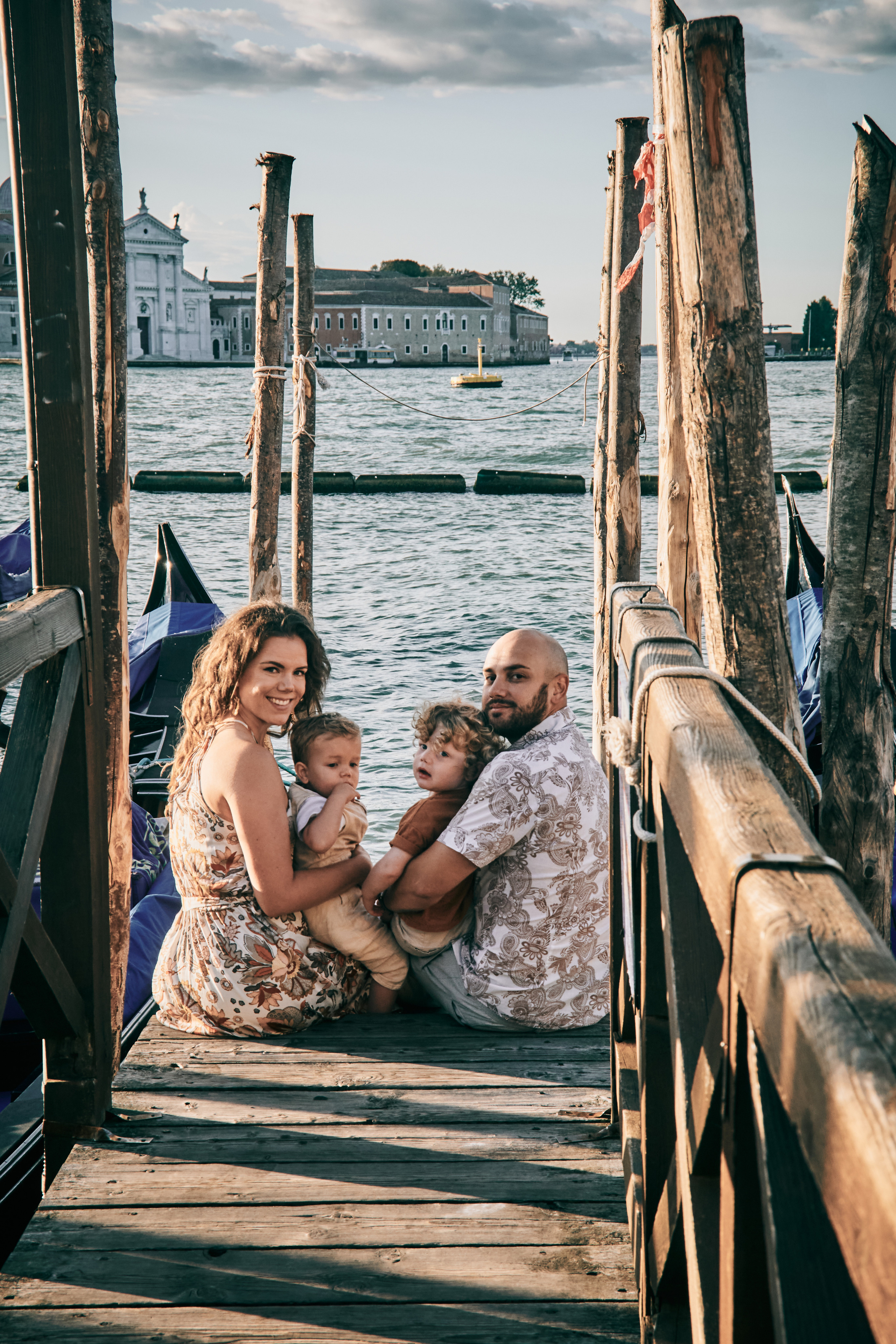 A happy family of four stands in front of the iconic St. Mark's Square, with the beautiful Venetian architecture and gondolas in the background.