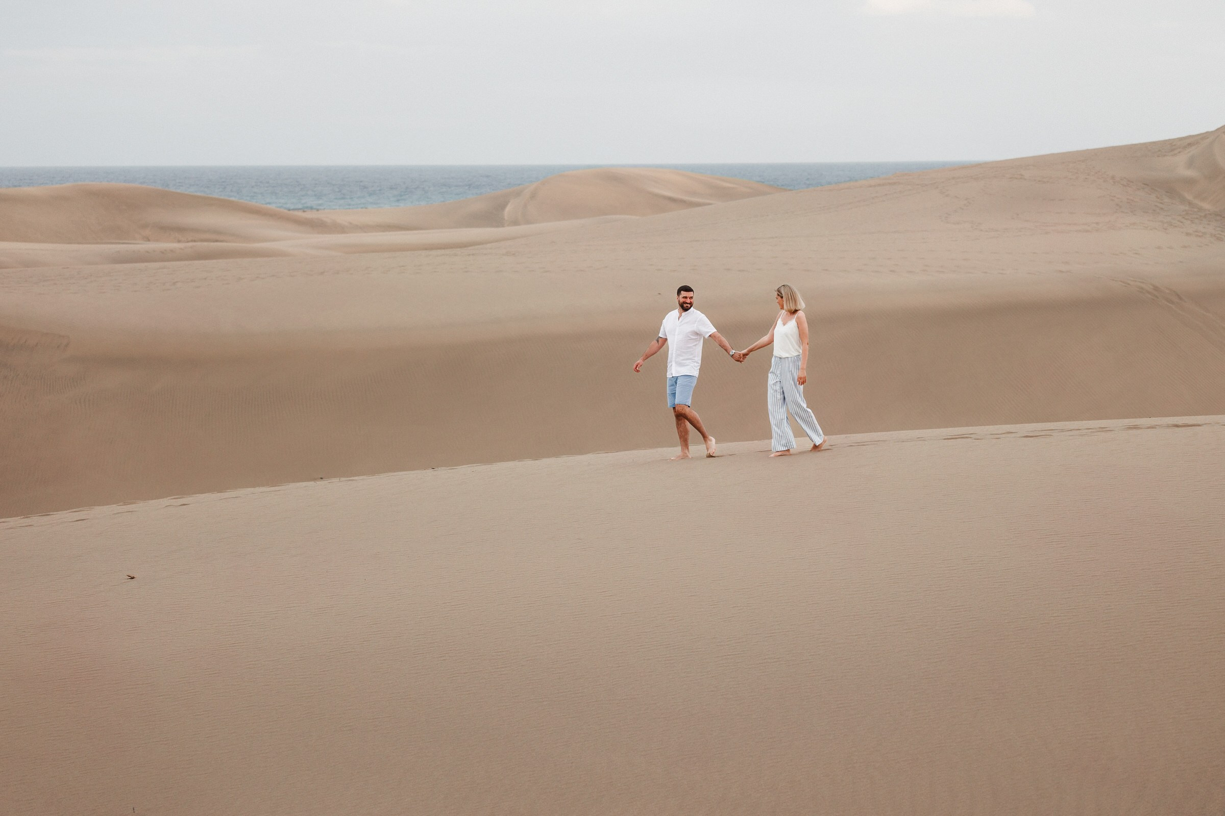 Photographer Gran Canaria - A couple in white attire holding hands while walking across a desert landscape with sand dunes and a distant ocean.