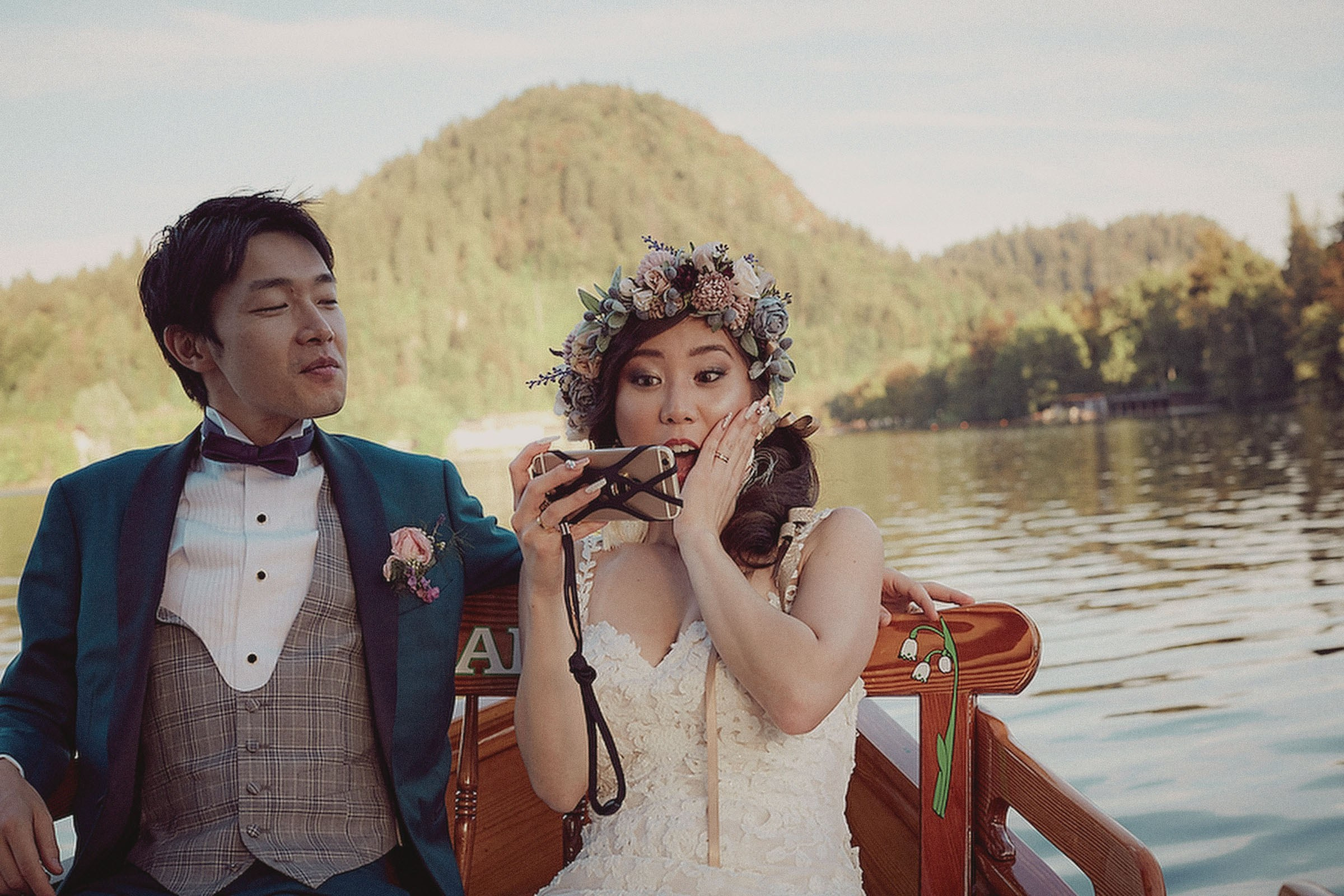A smiling groom watches as his bride mimics a surprised expression as she takes a selfie aboard a private boat.