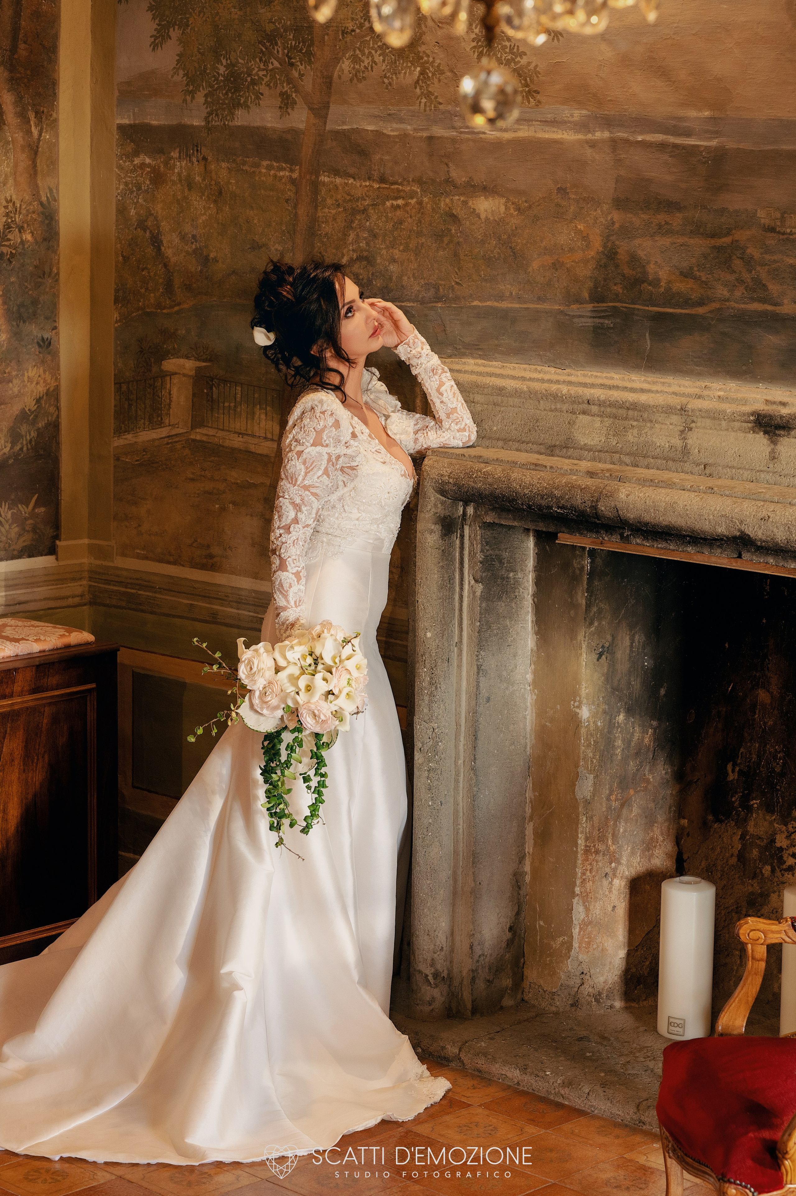 sposa pensierosa vicino al camino con lampadario e bouquet in mano