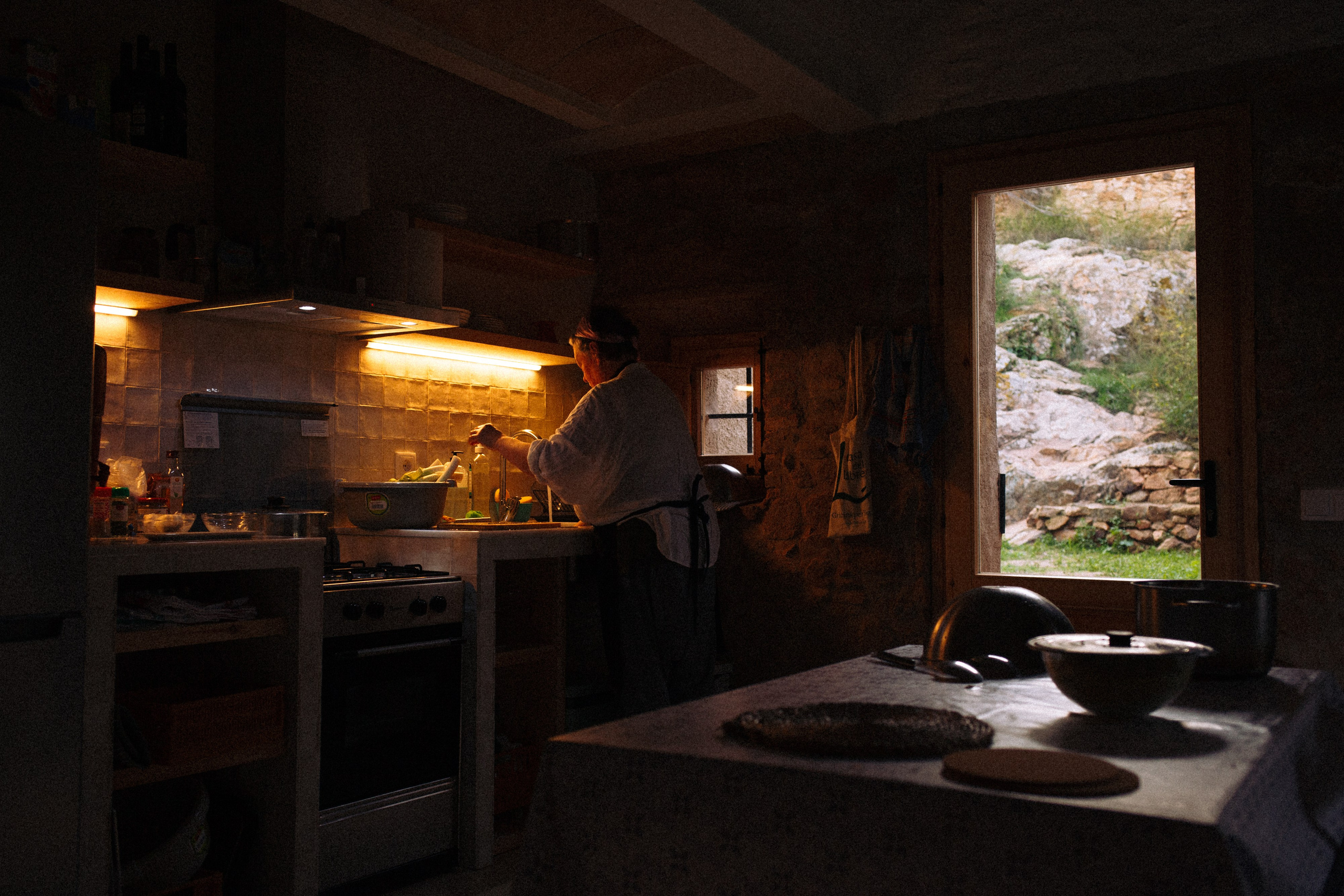 Intimate, personal atmosphere of cooking lunch in a dim room; through a window-door, part of a street and historic fortress are visible. A woman chef prepares food in warm light, creating a cozy scene
