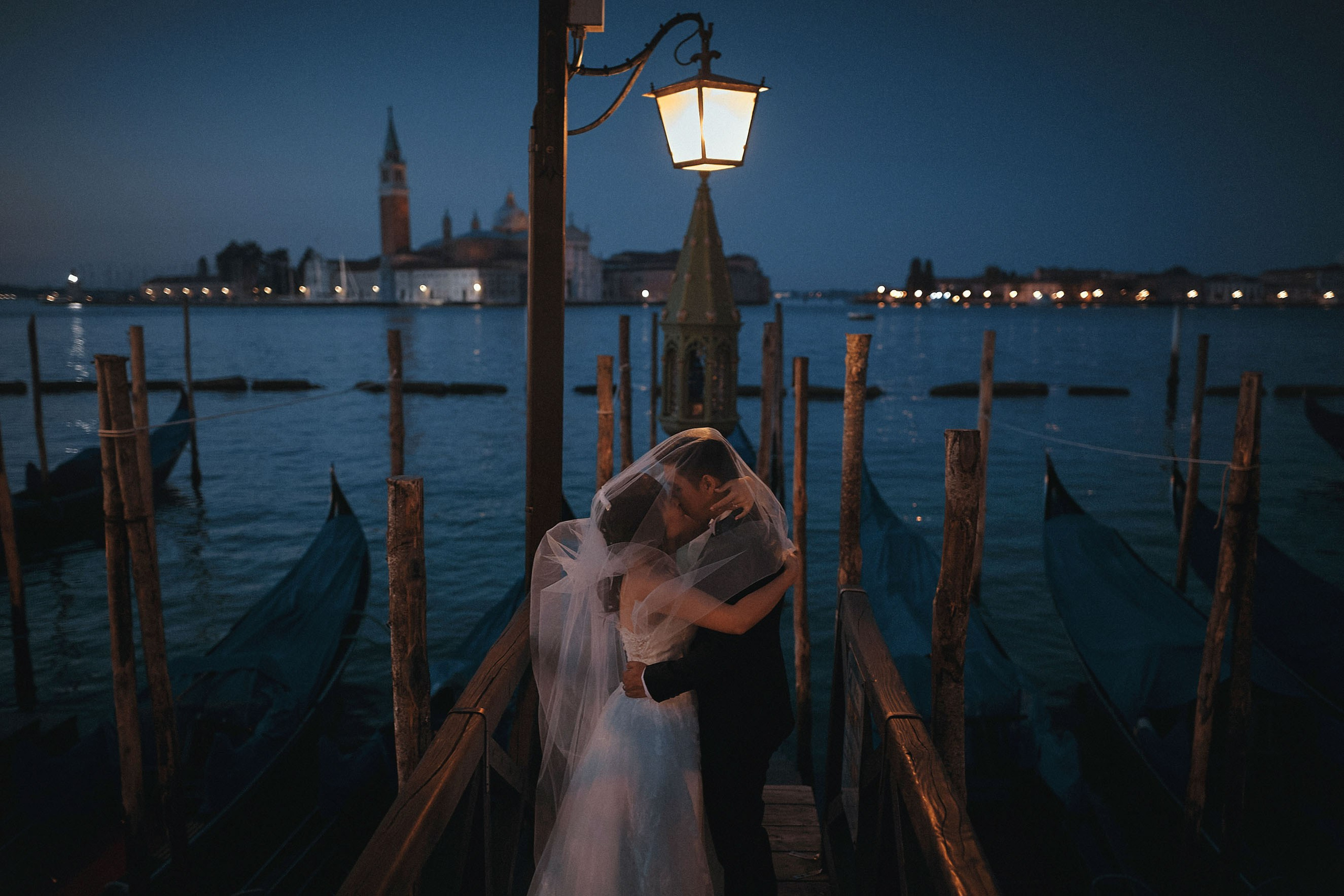 Romantic moment of a bride and groom kissing under a historic lantern, overlooking gondolas near San Marco Square, Venice, in the soft light of early morning before sunset.
