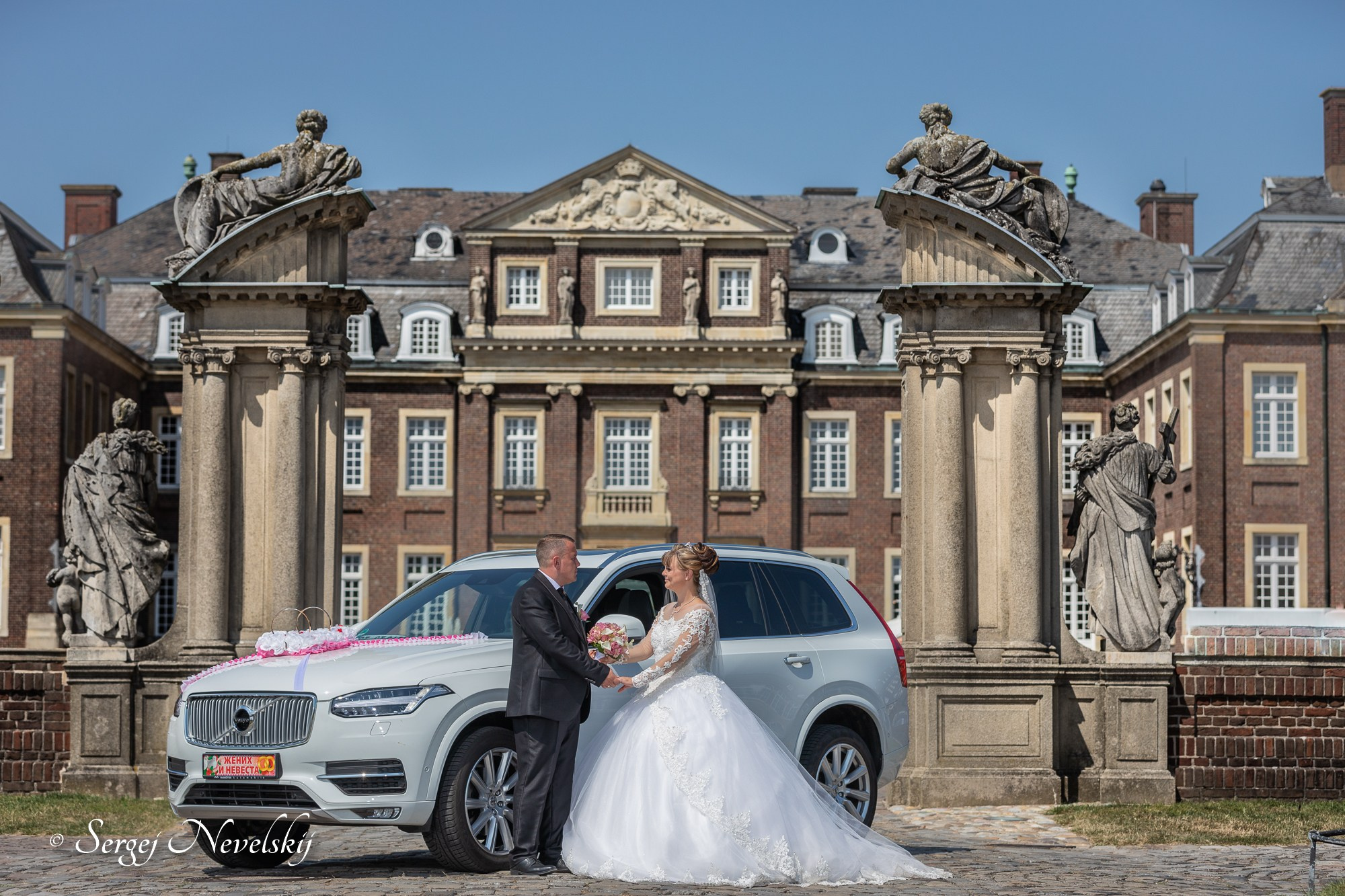 English:Newlyweds standing hand-in-hand in front of a grand historic palace with classical columns and statues. Bride in a luxurious princess-style white wedding dress with long lace sleeves and veil, groom in a classic black suit. White Volvo SUV wedding car decorated with pink floral garlands and “ЖЕНИХ И НЕВЕСТА” (Groom & Bride) license plate. Elegant and timeless wedding portrait under a clear blue sky. Photo by © Sergej NevelskijРусский:Молодожёны держатся за руки перед величественным историческим дворцом с колоннами и статуями. Невеста в роскошном свадебном платье принцессы с длинными кружевными рукавами и фатой, жених в классическом чёрном костюме. Белый Volvo, украшенный розовыми цветочными гирляндами и номером «ЖЕНИХ И НЕВЕСТА». Элегантный и вечный свадебный портрет на фоне ясного голубого неба. Фото © Sergej NevelskijDeutsch:Brautpaar Hand in Hand vor einem prächtigen historischen Schloss mit Säulen und Statuen. Braut im märchenhaften Prinzessinnen-Brautkleid mit langen Spitzenärmeln und Schleier, Bräutigam im klassischen schwarzen Anzug. Weißer Volvo als Hochzeitsauto mit rosa Blumengirlanden und dem Kennzeichen „ЖЕНИХ И НЕВЕСТА“ (Bräutigam & Braut). Elegantes und zeitloses Hochzeitsporträt unter klarem blauen Himmel. Foto © Sergej Nevelskij