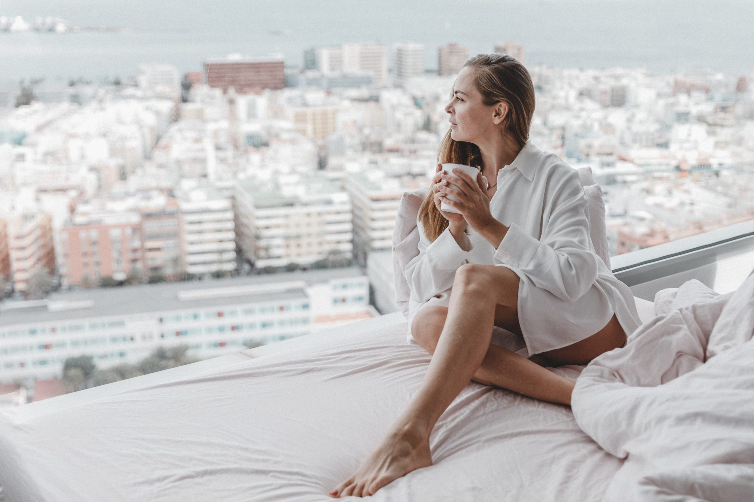 A young woman is sitting on a bed with a cup of coffee.