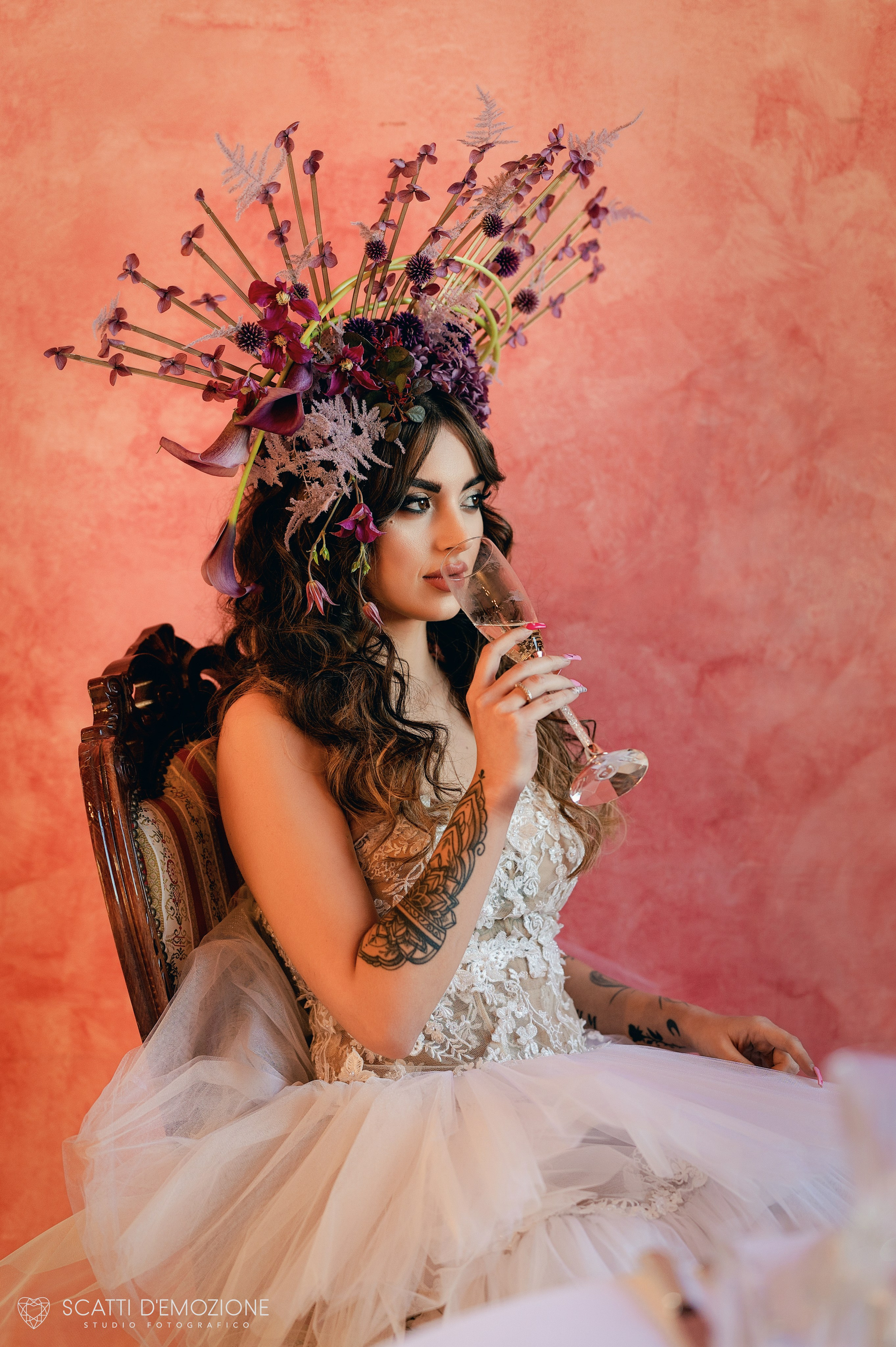 Sposa con acconciatura floreale in toni rosso e rosa indossa un abito da sposa in pizzo con schiena scoperta durante uno shooting di matrimonio artistico mentre sorseggia vino dal bicchiere.