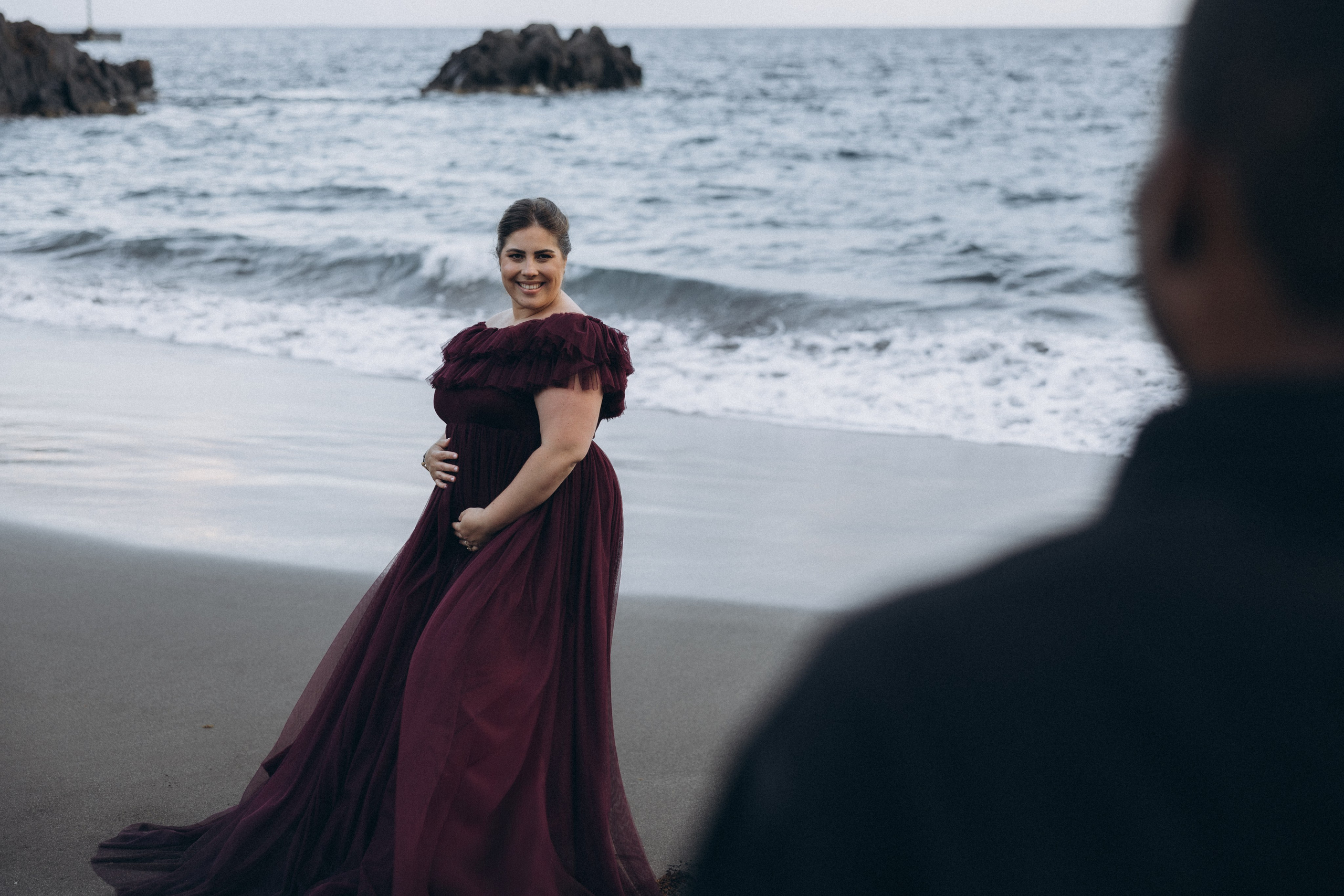 A glowing expectant mother standing on a cliff overlooking the ocean in Madeira, her dress flowing gently in the wind as the golden sunset casts a warm glow.