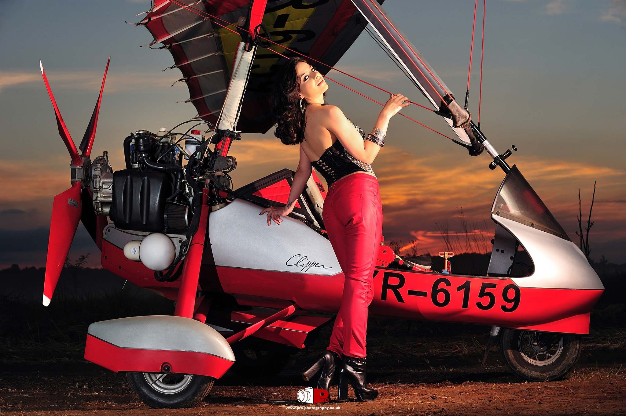 A woman in red pants and a black corset posing with a vintage ultralight aircraft during sunset.