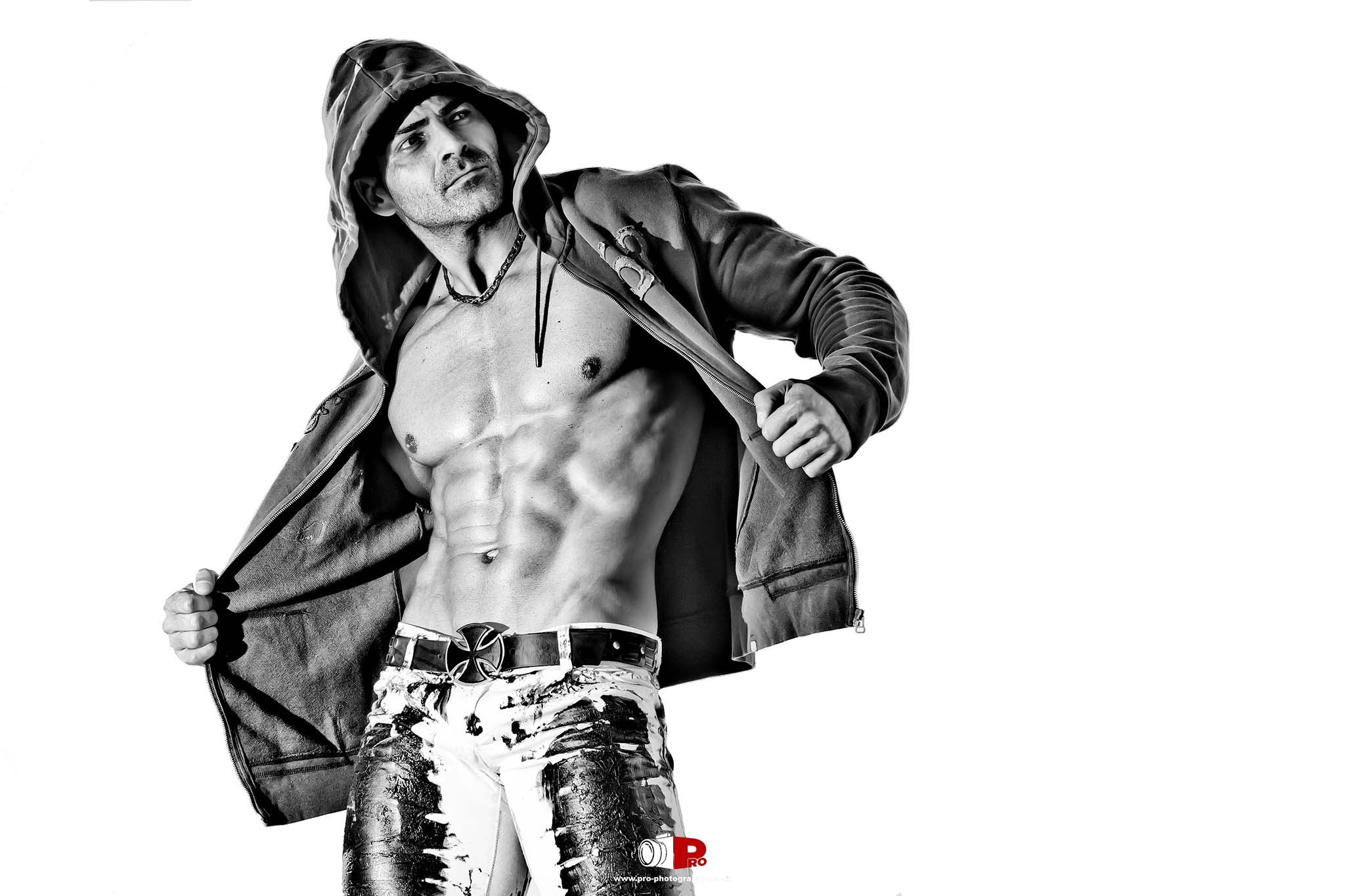 Male fitness model in a hoodie revealing sculpted abs with a confident pose against a white background.