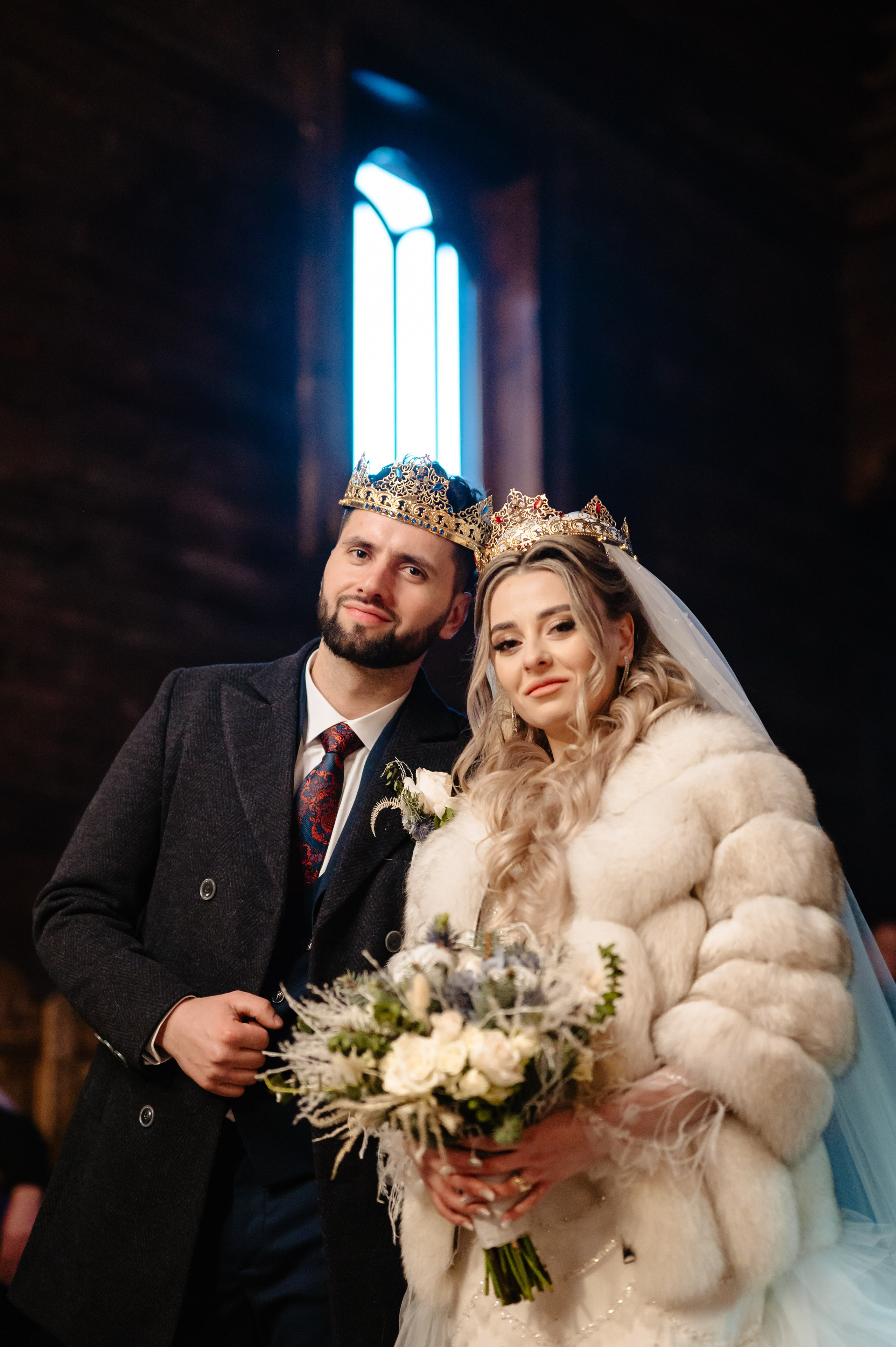 Mirele și mireasa, purtând coroane tradiționale, într-un moment solemn și plin de emoție, în interiorul unei biserici de lemn. Eleganță și tradiție — mireasa în blană albă și mirele în costum clasic, încununați în cadrul ceremoniei religioase. Moment de poveste: mireasa și mirele, împodobiți cu coroane aurii, într-o atmosferă caldă