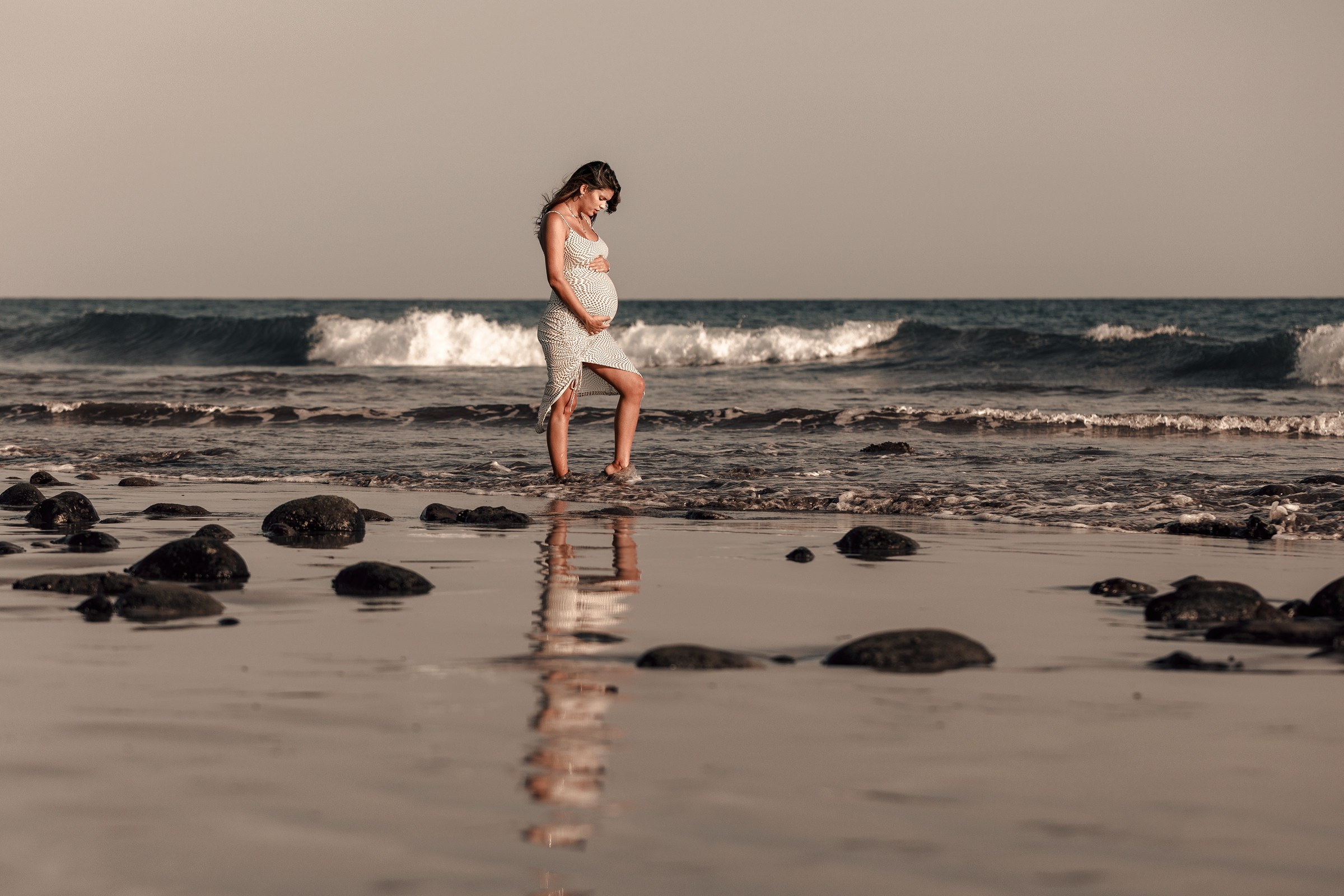 A pregnant woman standing on the beach in front of rocks.