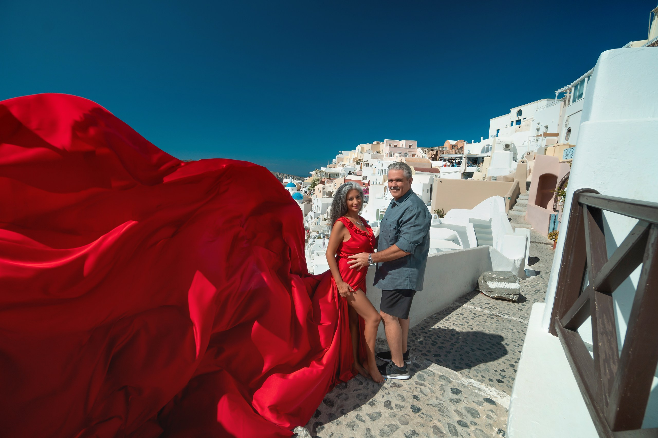 Elegant plus size model in red gown with V-neckline and ruffles, against colorful Cycladic architecture in Santorini.