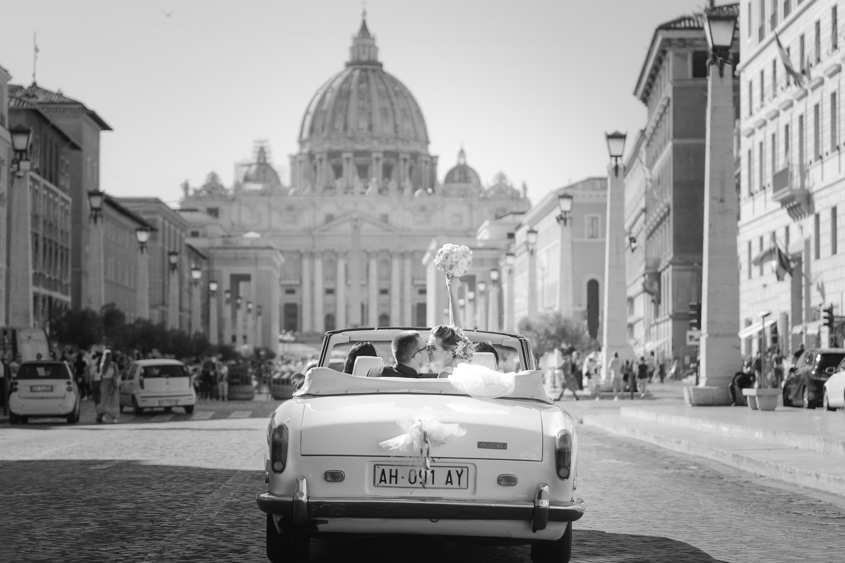 Sposi in fuga felice davanti al Colosseo, con lo sposo in smoking e la sposa in un fluttuante abito bianco, catturati in una fotografia in bianco e nero che esprime gioia e movimento nel cuore di Roma