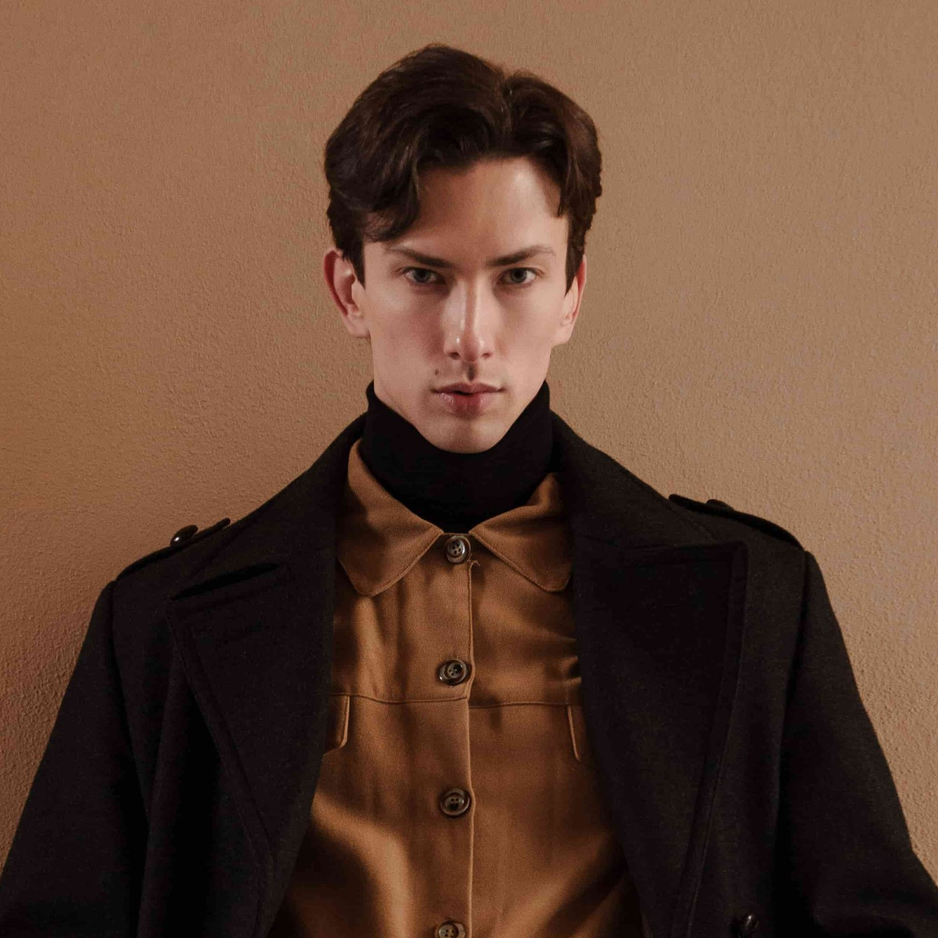 Male model in black coat and brown shirt leaning against wall