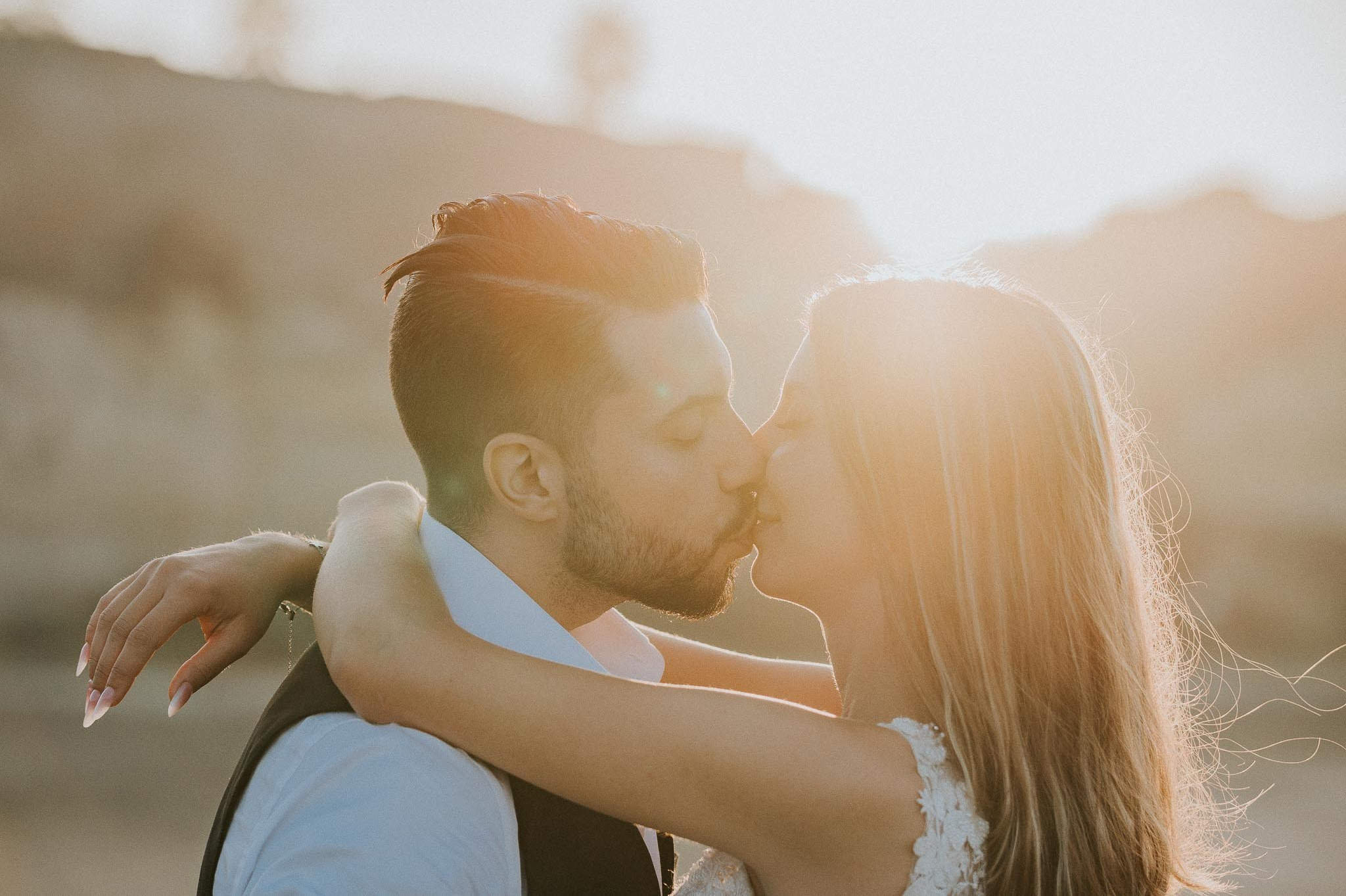 sposi si scambiano un tenero bacio nel calore del tramonto, con lui in un elegante gilet e lei in un abito da sposa decorato, catturando un momento di intima felicità.