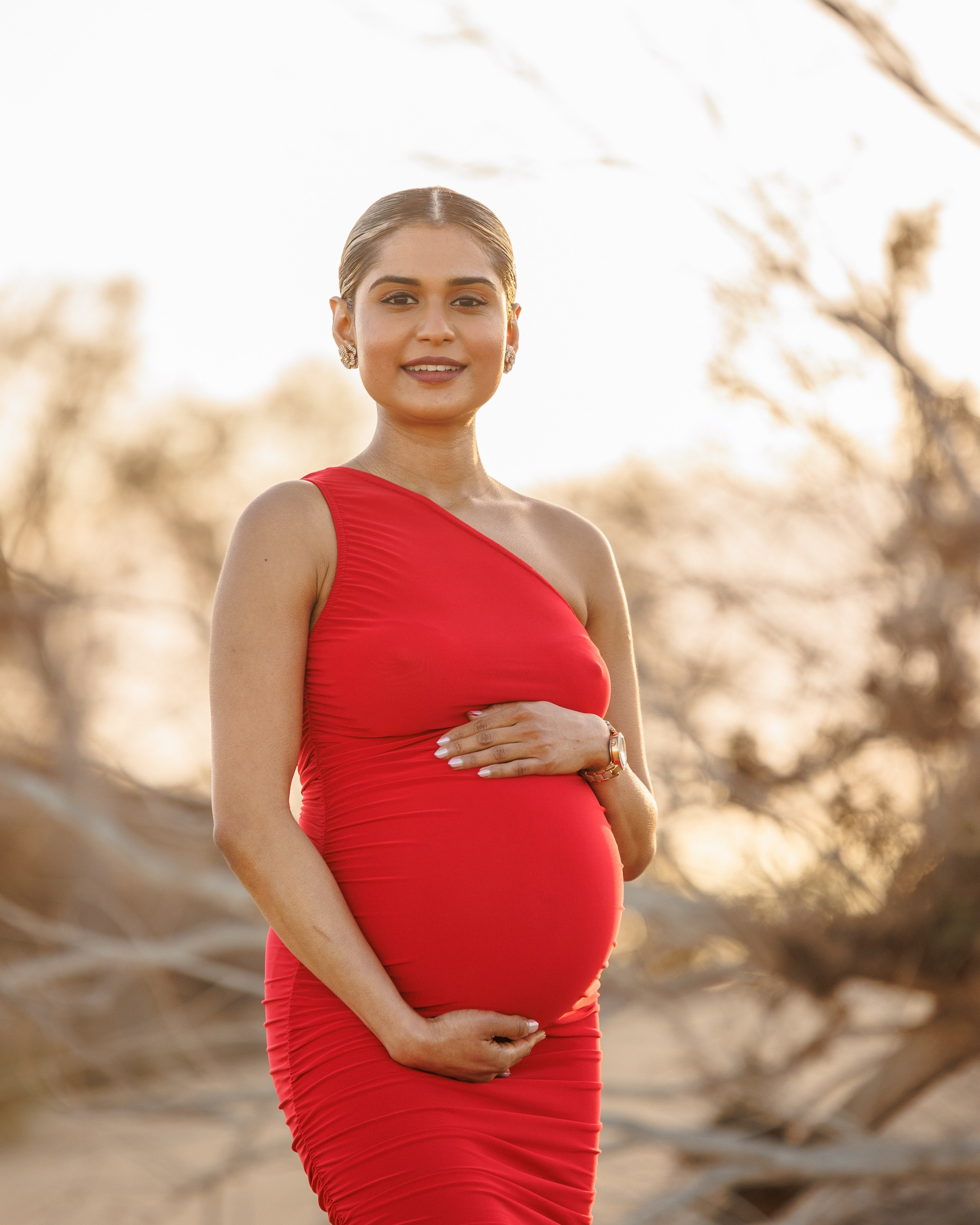 Meloneras, Grande Canarie. Une femme enceinte vêtue d'une robe rouge asymétrique se tient dehors, tenant son ventre à deux mains et souriant à l'objectif.