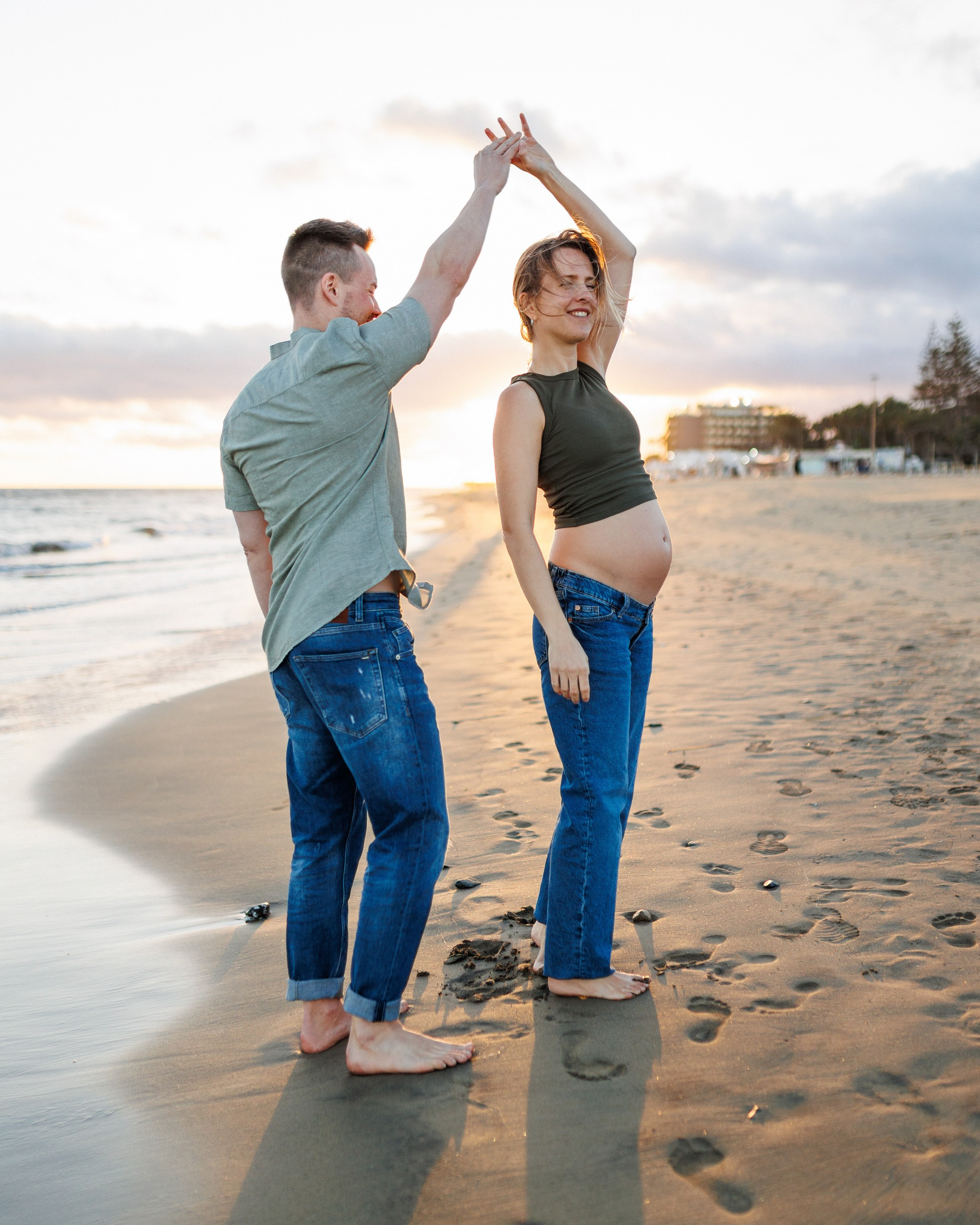 Maspalomas, Grande Canarie. Un couple pieds nus danse sur une plage de sable au coucher du soleil. La femme est visiblement enceinte, et tous deux sont vêtus de façon décontractée : jeans et chemise.