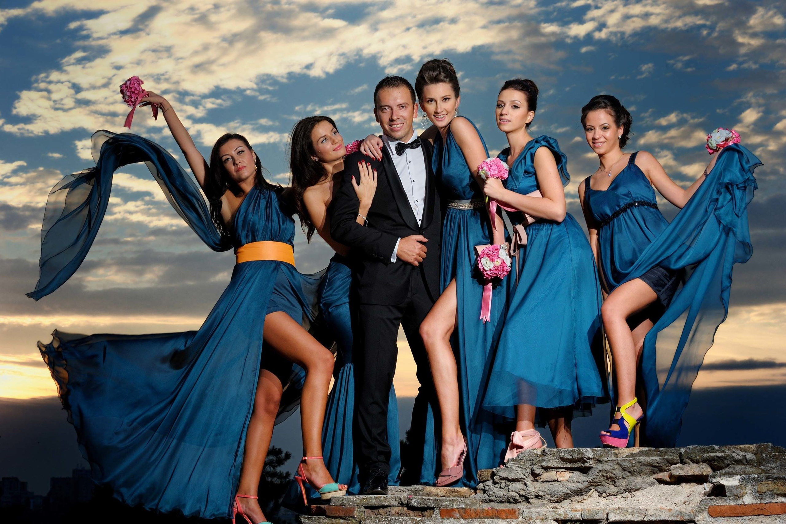 Groom surrounded by vibrant bridesmaids in flowing blue dresses under dramatic sky