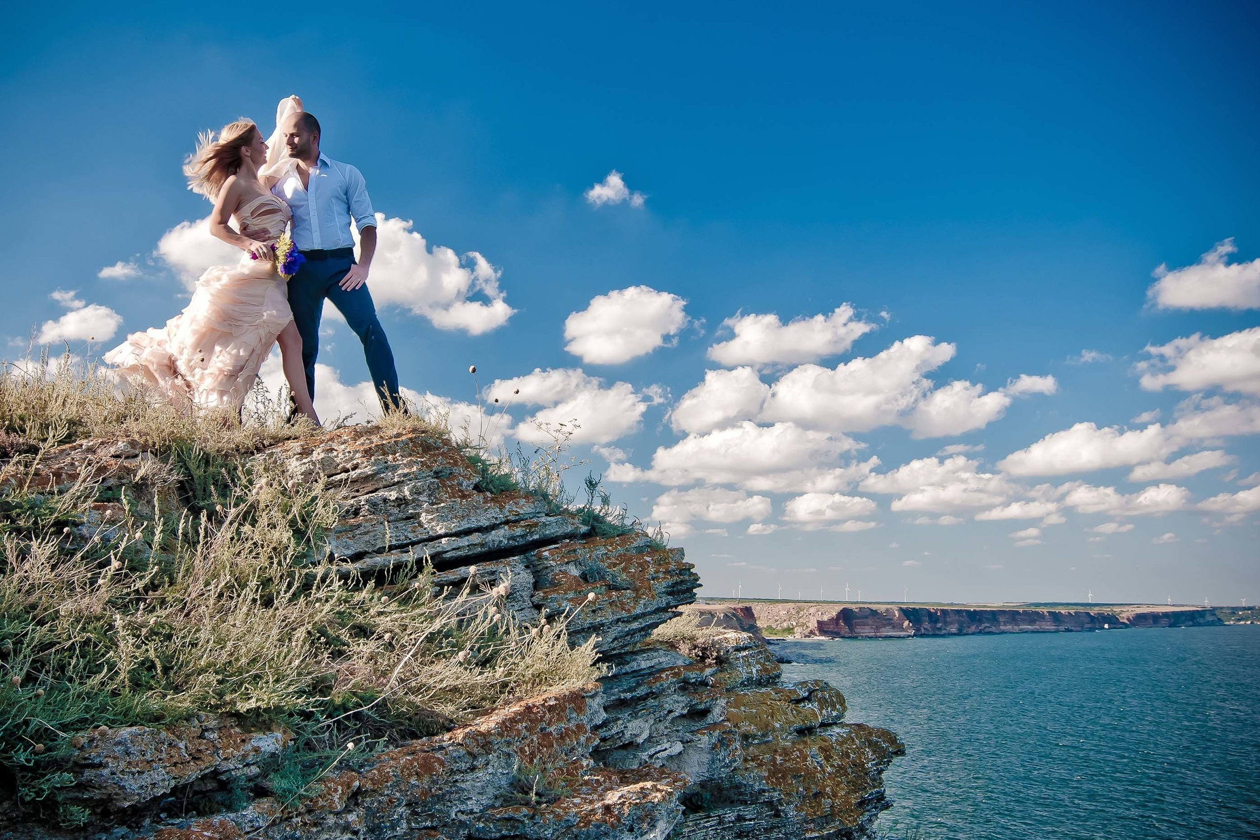 Bride and groom standing on a cliff edge overlooking the sea with blue sky and clouds, Kaliakra, Bulgaria