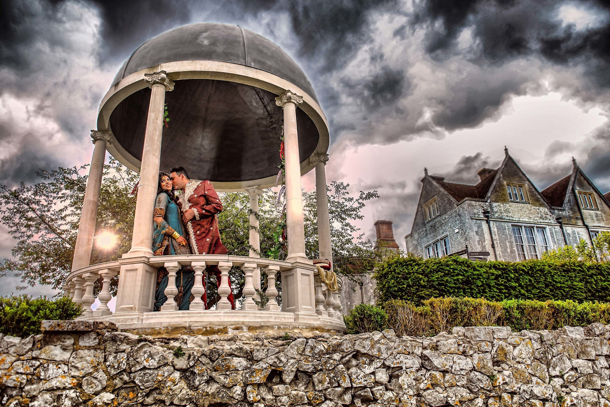 Asian wedding photo session with groom whispering at his bride under a dramatic sky at Froyle Park, Hampshire, UK Bride and groom in traditional wedding attire sharing a kiss in a pavilion with stormy sky