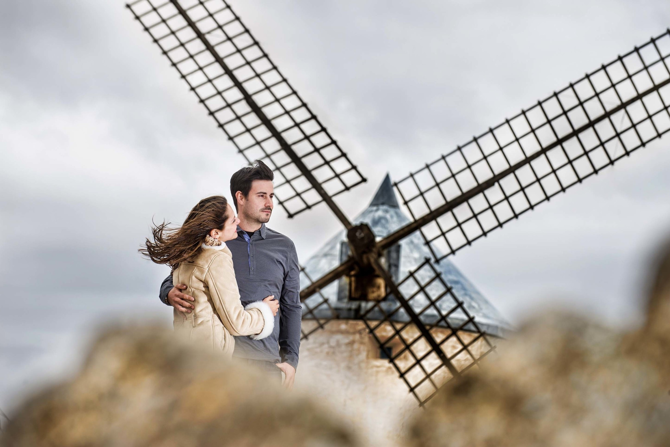 A couple sharing a moment in front of a historic windmill under cloudy skies in Toledo, Spain