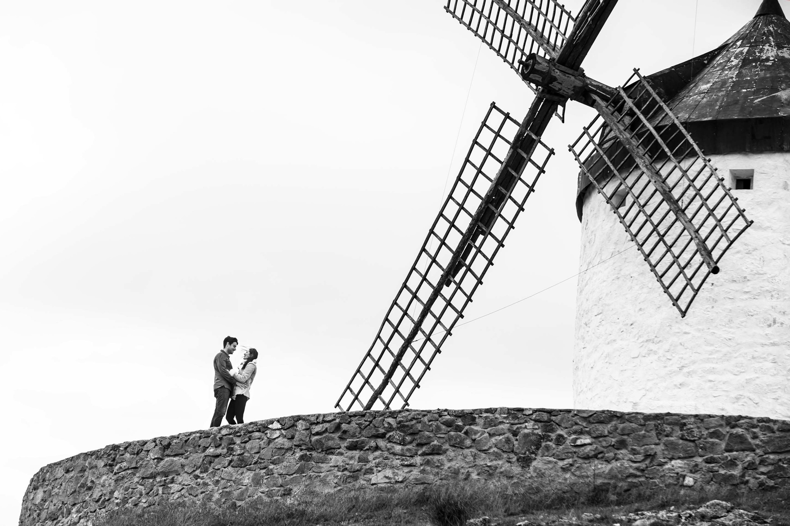 Black and white photo of a couple embracing in front of an old windmill on a stone platform in Toledo, Spain
