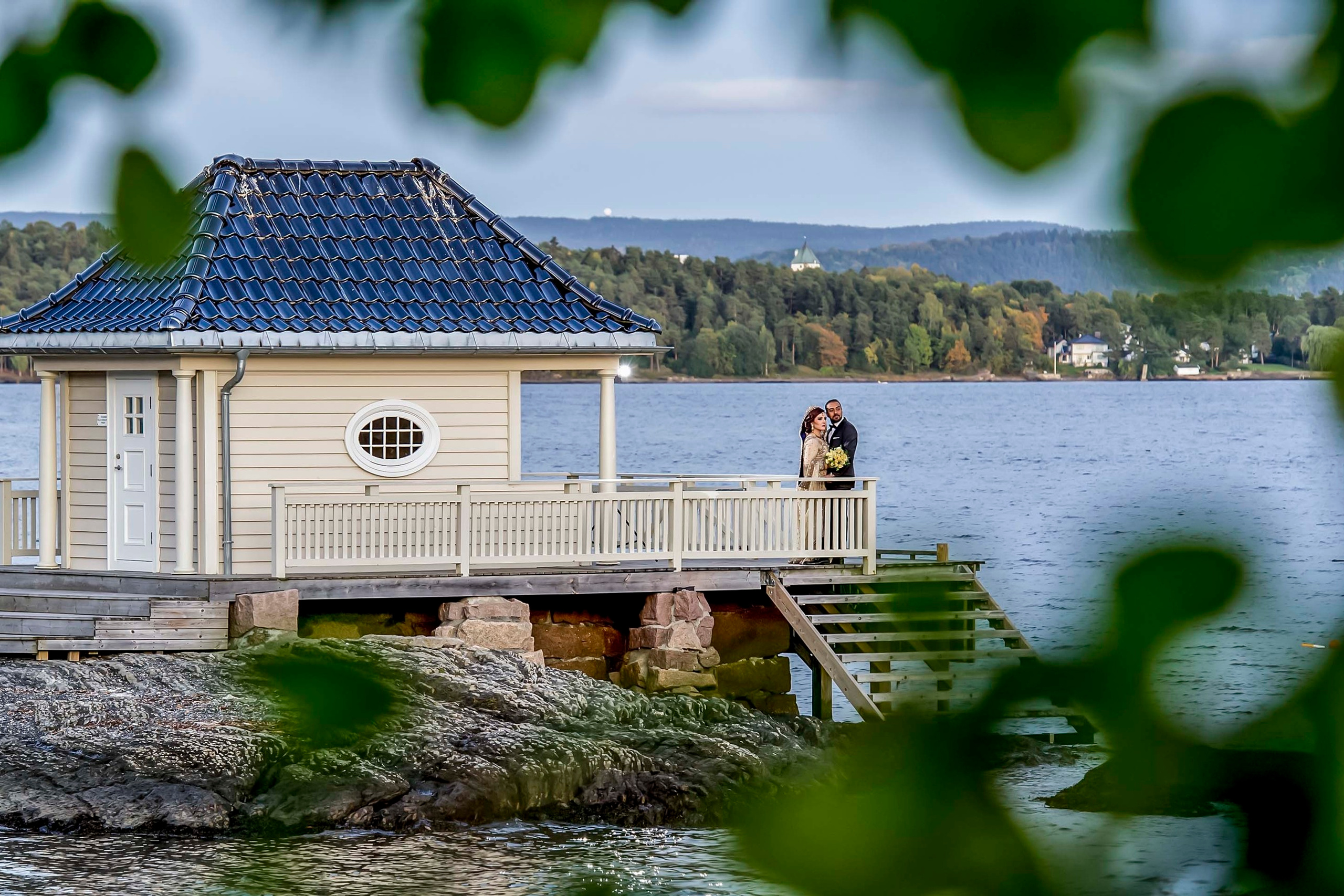Asian wedding photo session at the shore near Scandic Fornebu in Oslo, Norway. Bride and groom standing on a wooden deck by a lake surrounded by nature.