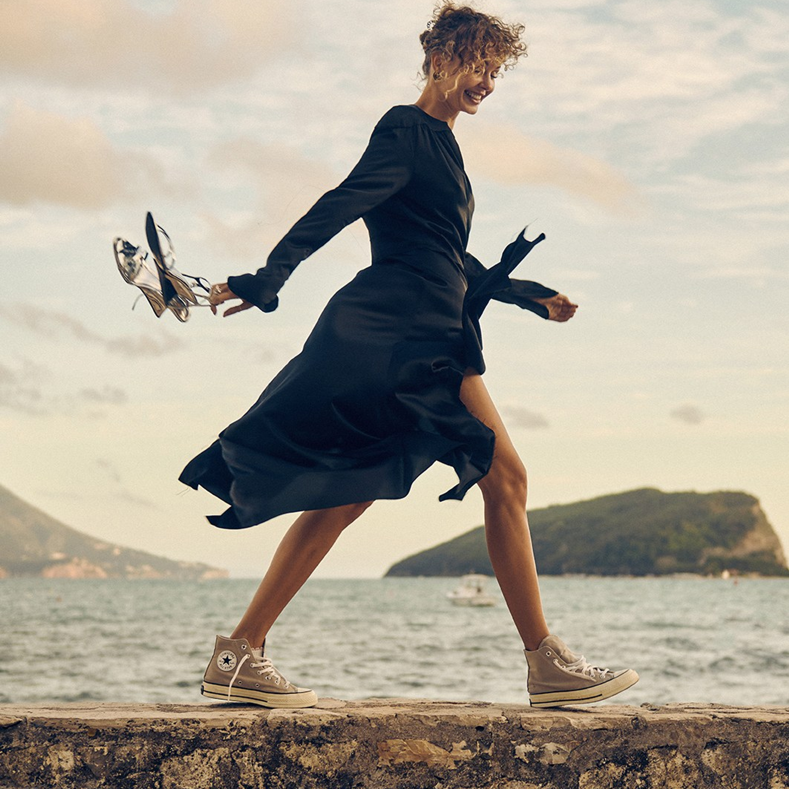 Artistic outdoor photoshoot with a curly haired model wearing elegant black dress, captured in natural light by the sea