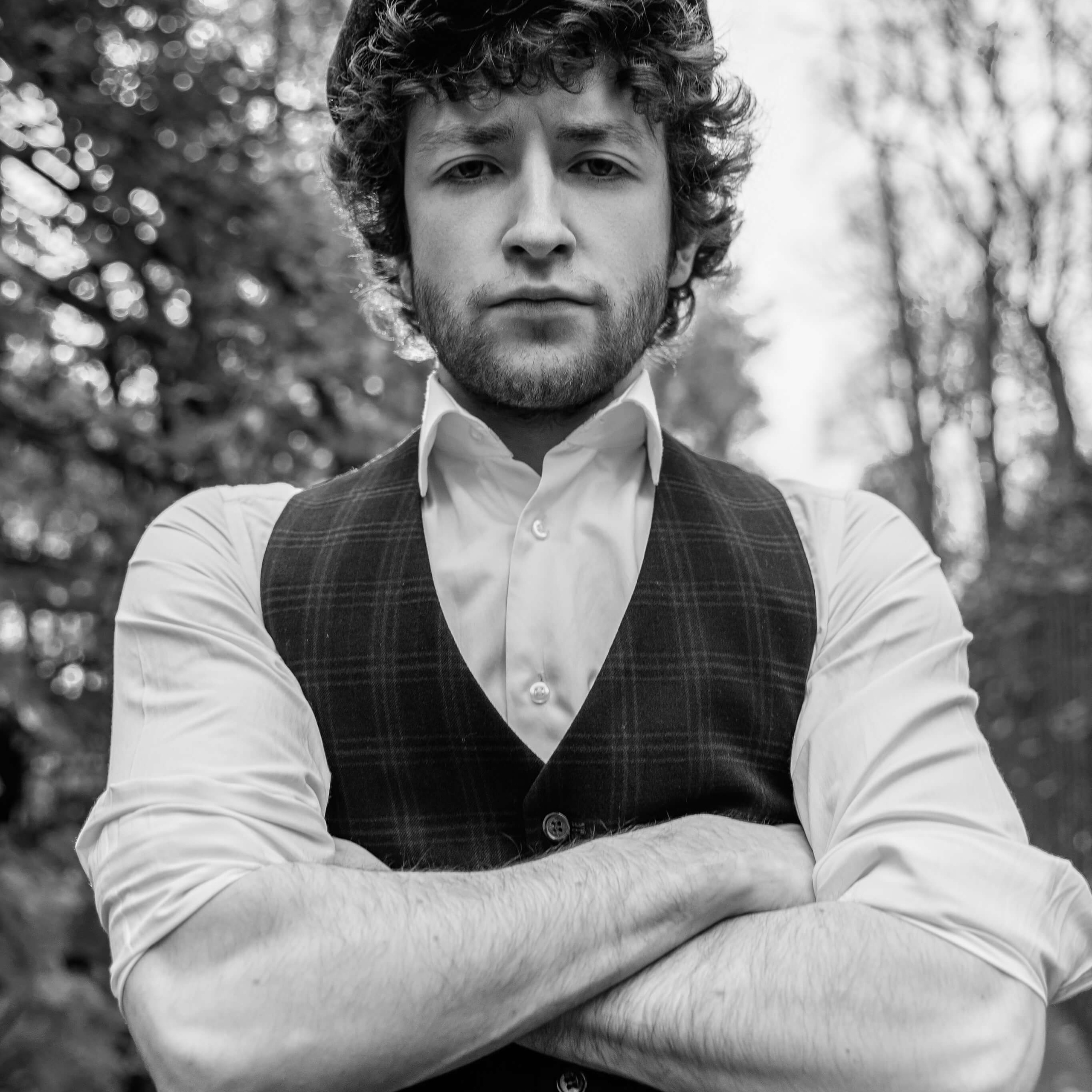 A black and white portrait of a man in vintage attire, with crossed arms and a confident stance, wearing a flat cap and a waistcoat