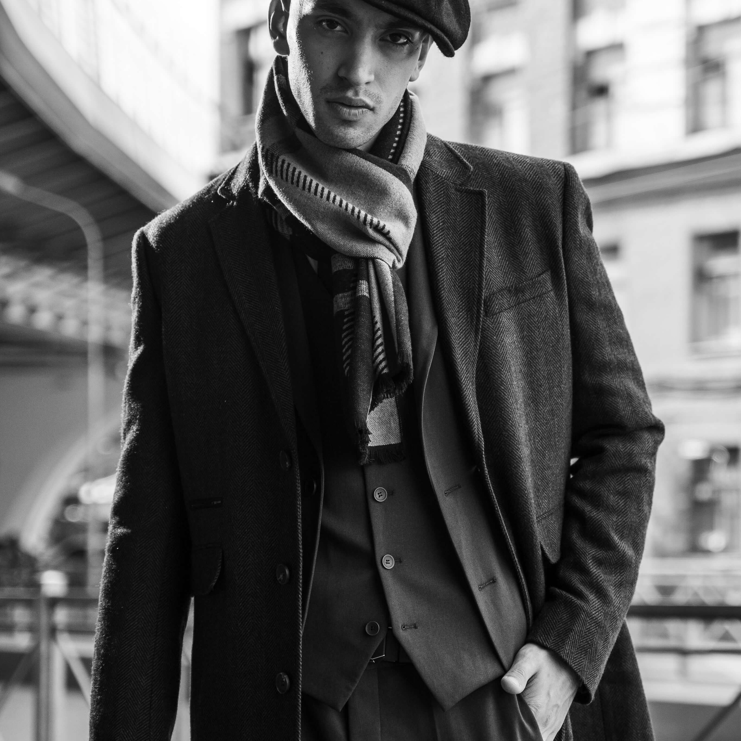 A monochromatic image of a fashionably dressed person in a textured coat, scarf, and flat cap, exuding a classic and timeless style, standing confidently with an urban landscape in the background