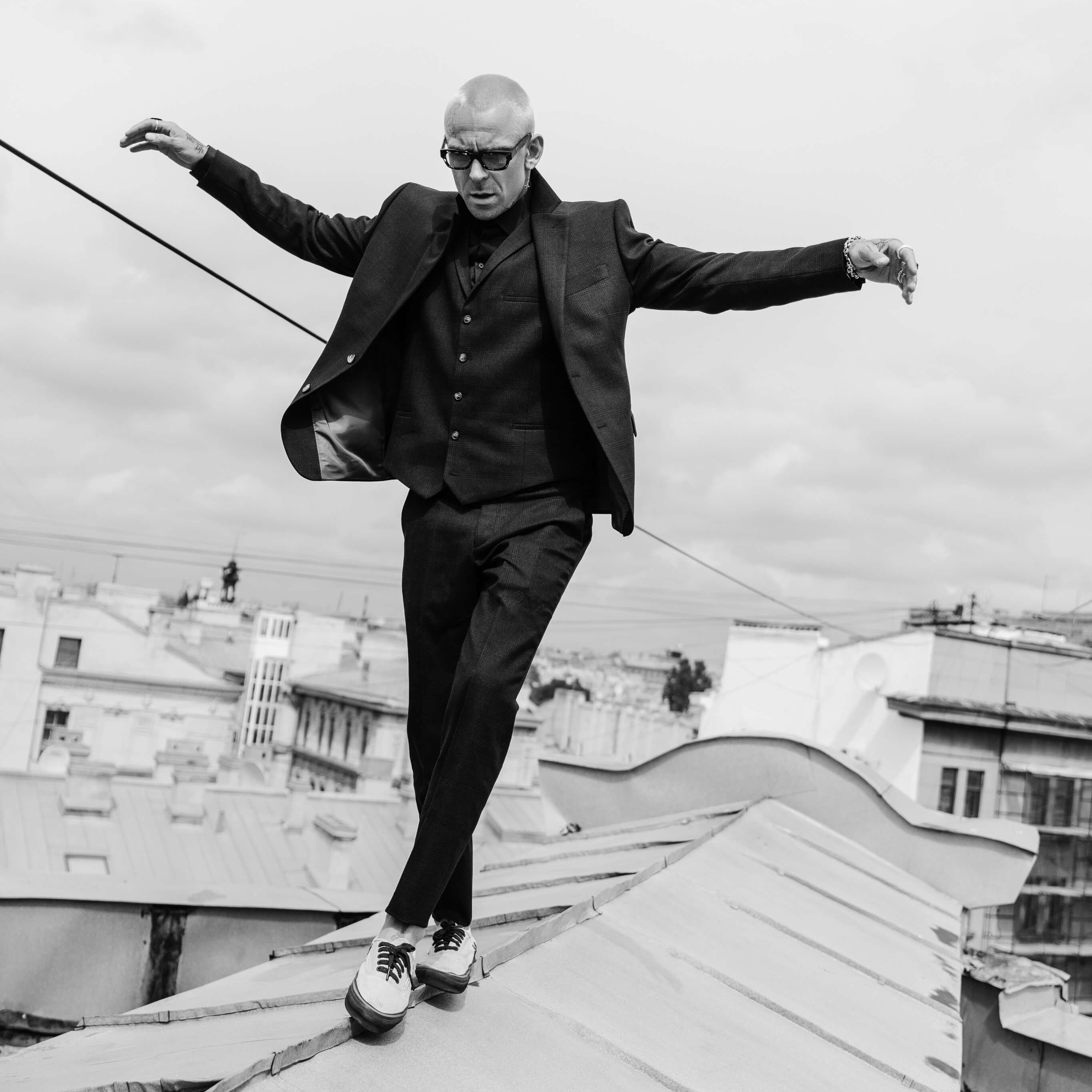 A black and white photograph captures a man balancing on a rooftop, his posture focused and deliberate, against the backdrop of a sprawling cityscape, conveying a sense of adventure and urban exploration