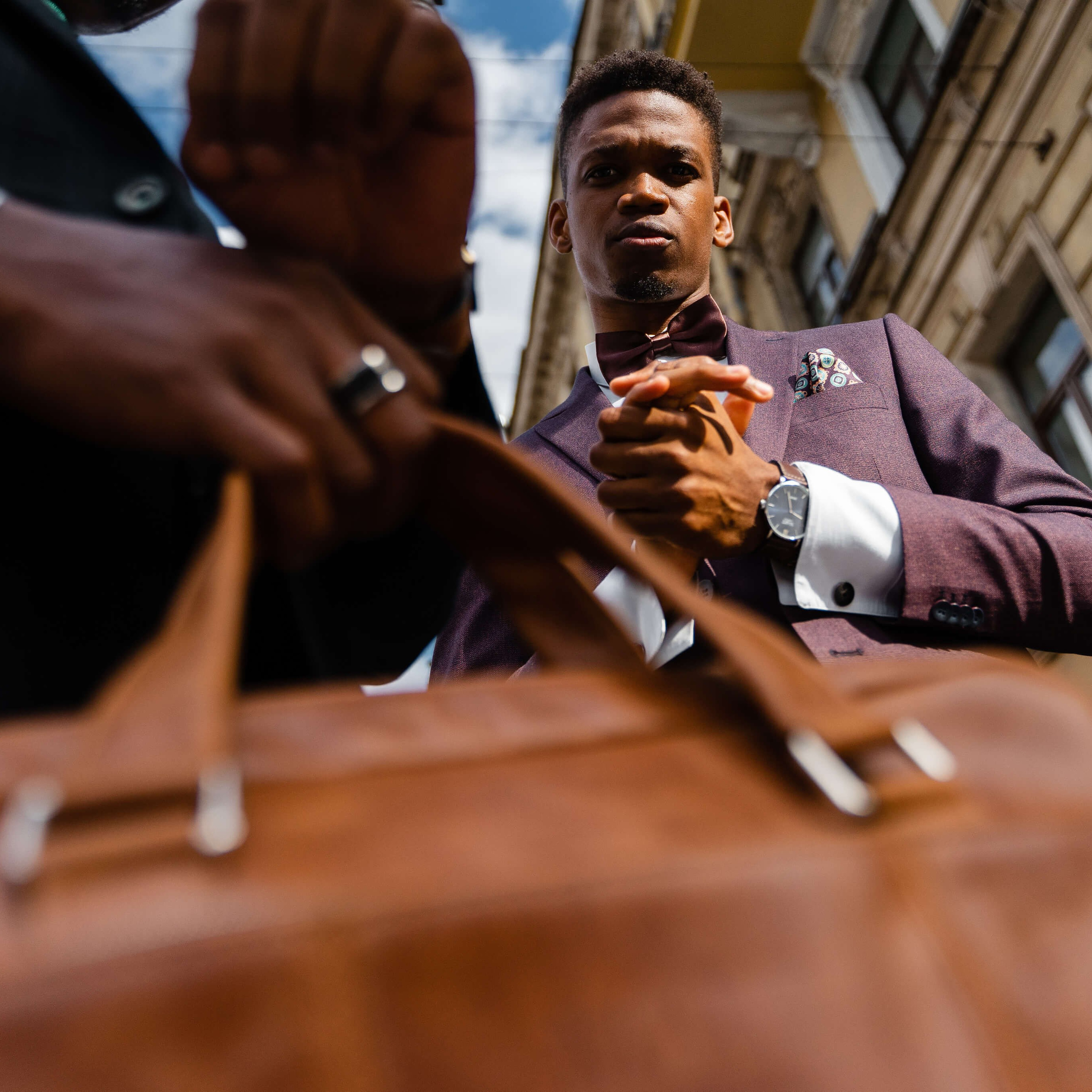 A ground-level perspective of a suave man in a tailored suit, focusing on his elegant wristwatch and leather bag, set against the city's architecture