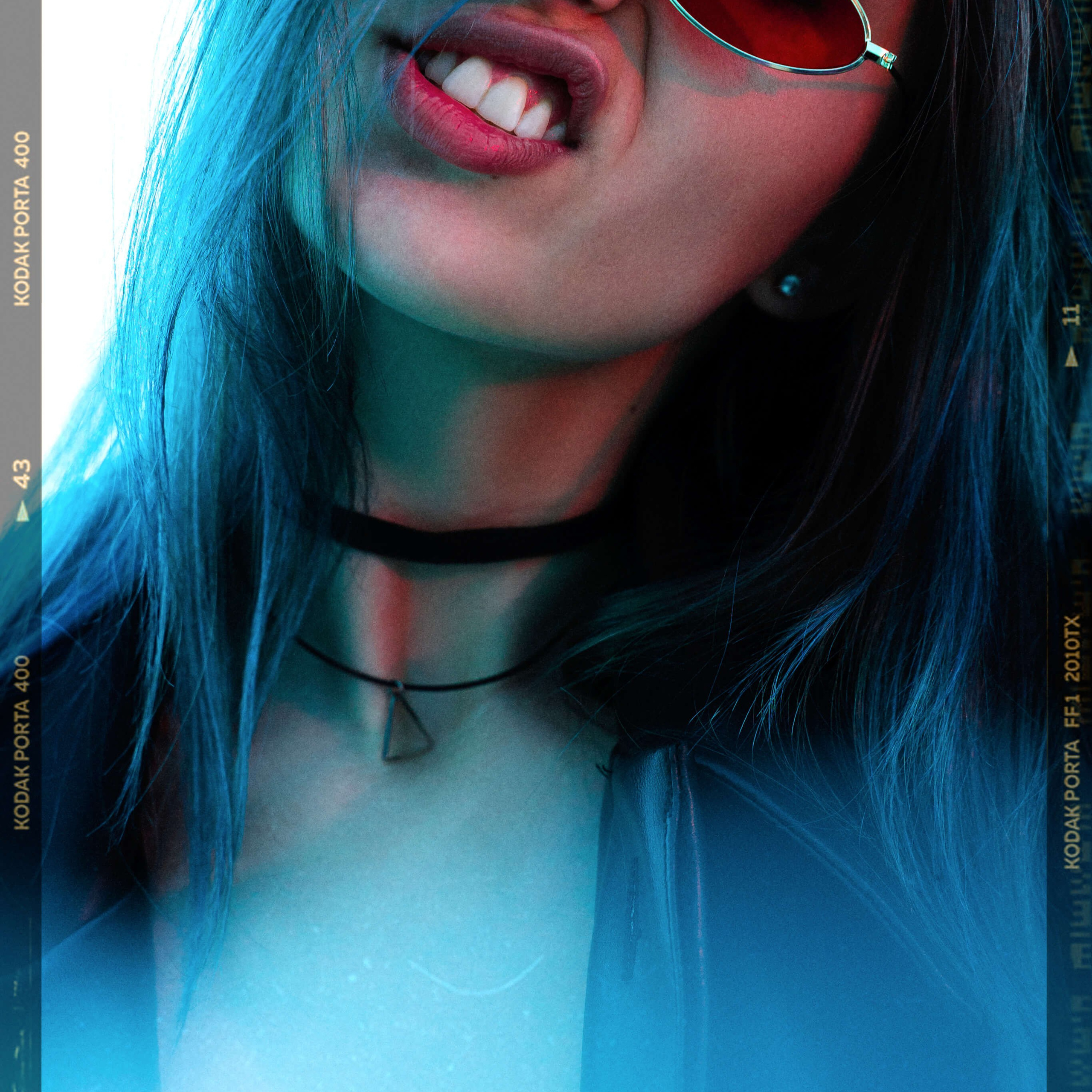 An artistic portrait of a person with vibrant blue hair, partially obscured by red-tinted aviator sunglasses, set against a backdrop of cool blue tones, with the frame mimicking the classic look of Kodak Portra 400 film