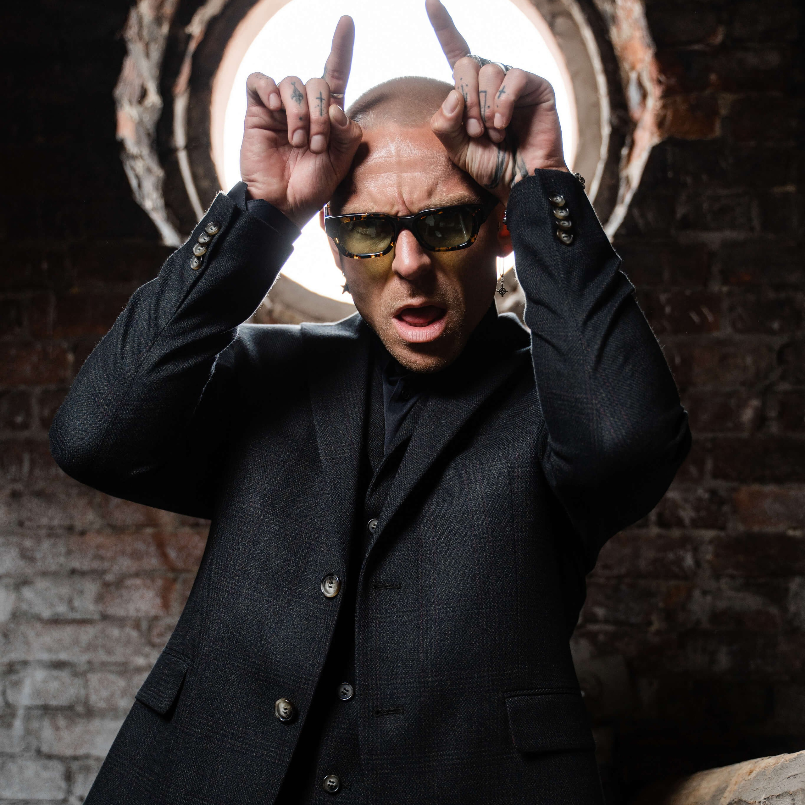 A man in a tailored suit and stylish sunglasses poses humorously with fingers on his head, mimicking horns, blending formal fashion with a touch of whimsy