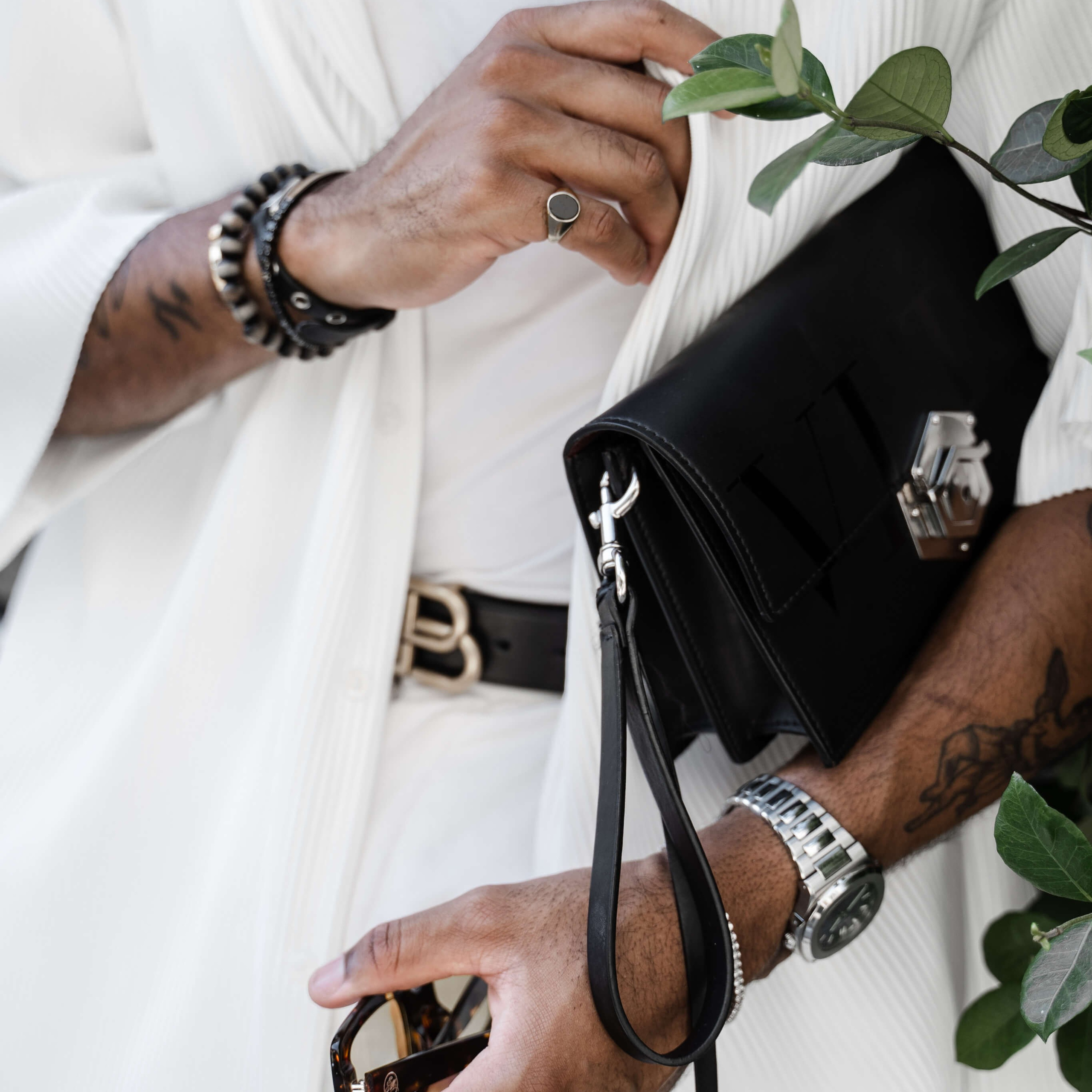 A person in a white pleated shirt holding a small black leather bag and sunglasses, with a silver watch and multiple bracelets on the wrist, showcasing a stylish and sophisticated fashion accessory arrangement